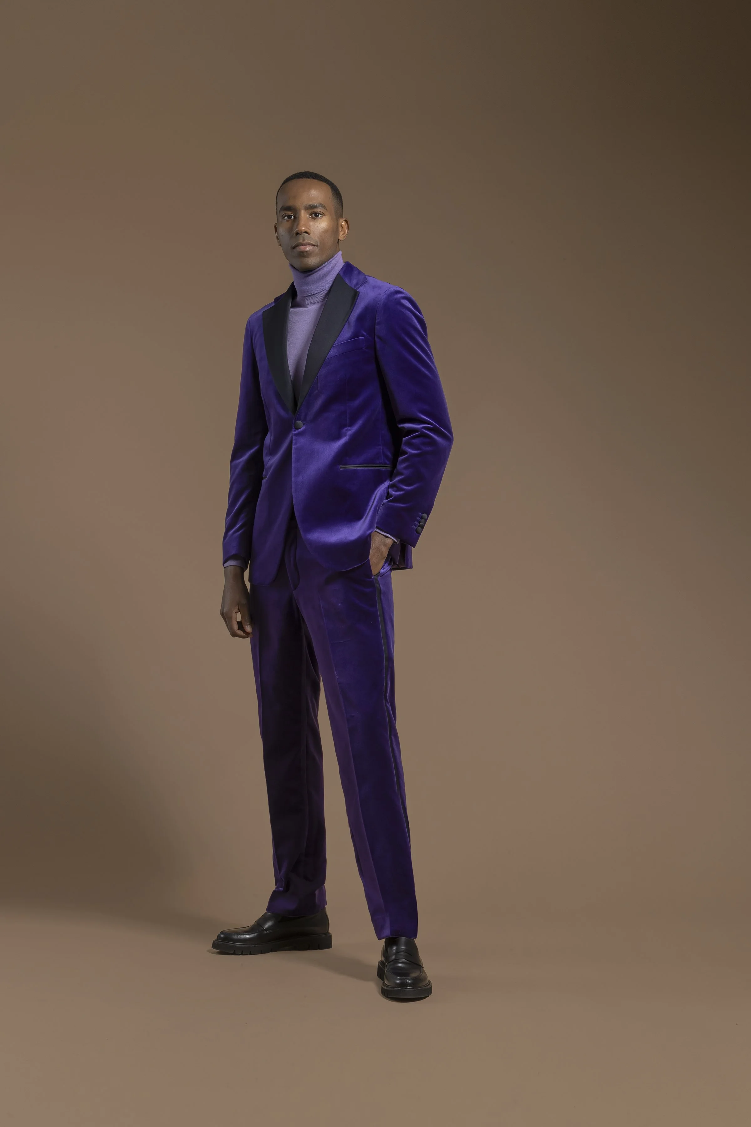 A man standing with his left hand in his pocket, wearing a Scabal purple velvet suit with black lapels, a purple turtleneck, and black shoes, against a plain brown background.