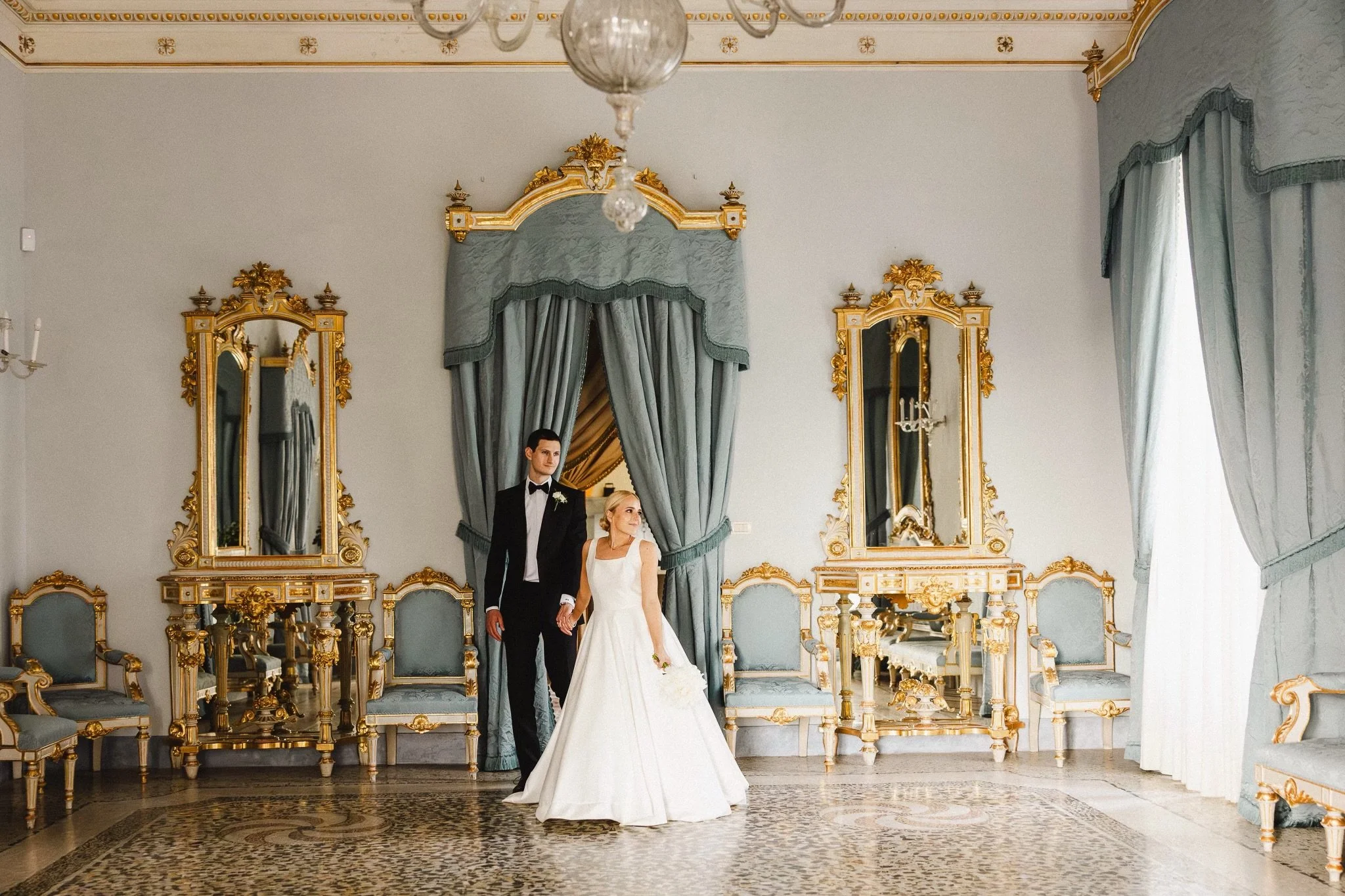 A bride and groom wearing Scarborough Custom Tailoring tuxedo standing hand in hand in an ornate, gold-accented room with tall mirrors and curtains.