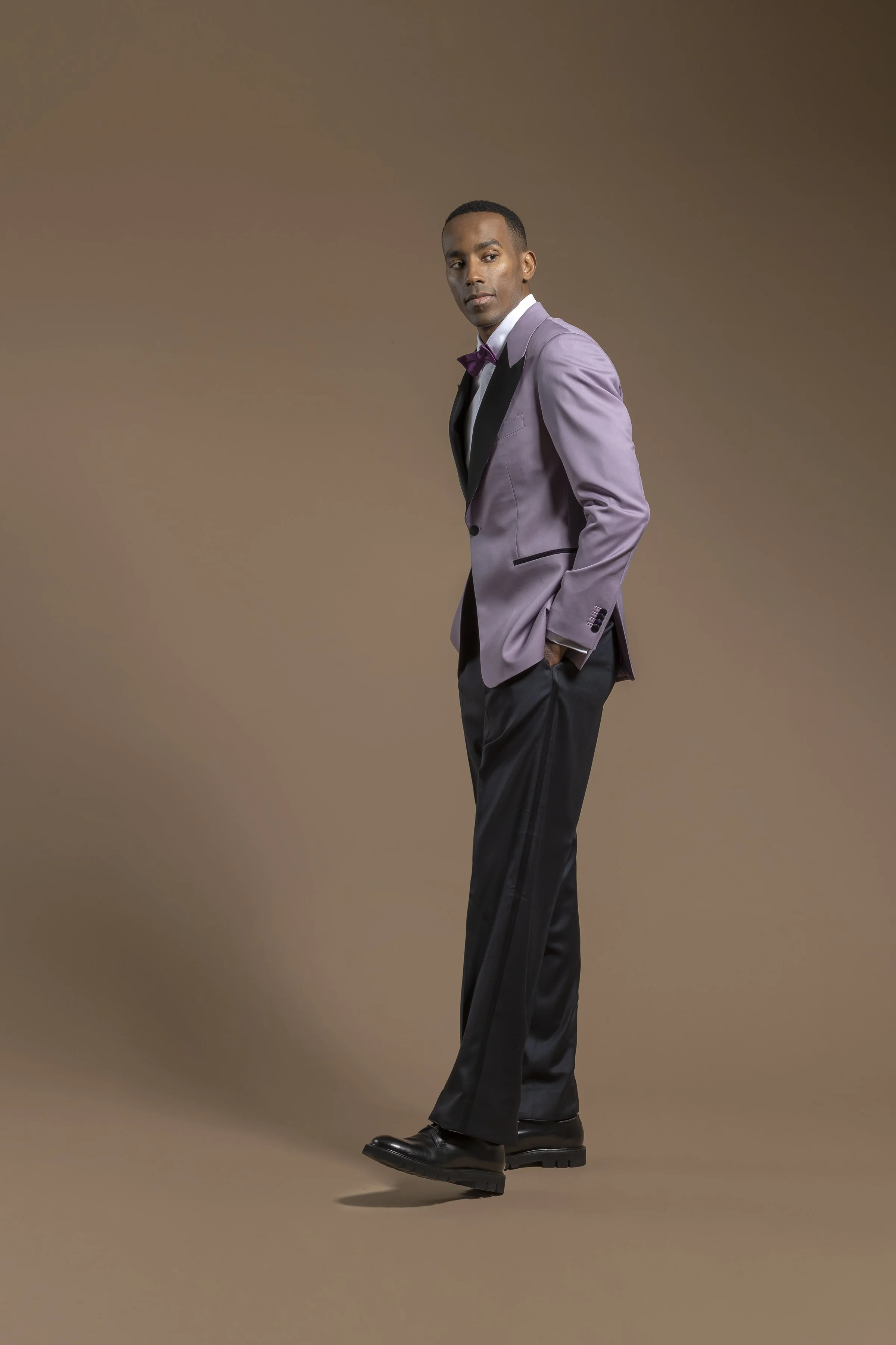 A man in a Scabal lavender suit with black lapel, black pants, and black shoes, wearing a white shirt and purple bow tie, stands against a plain brown background. He has his hands in his pockets and is looking at the camera.