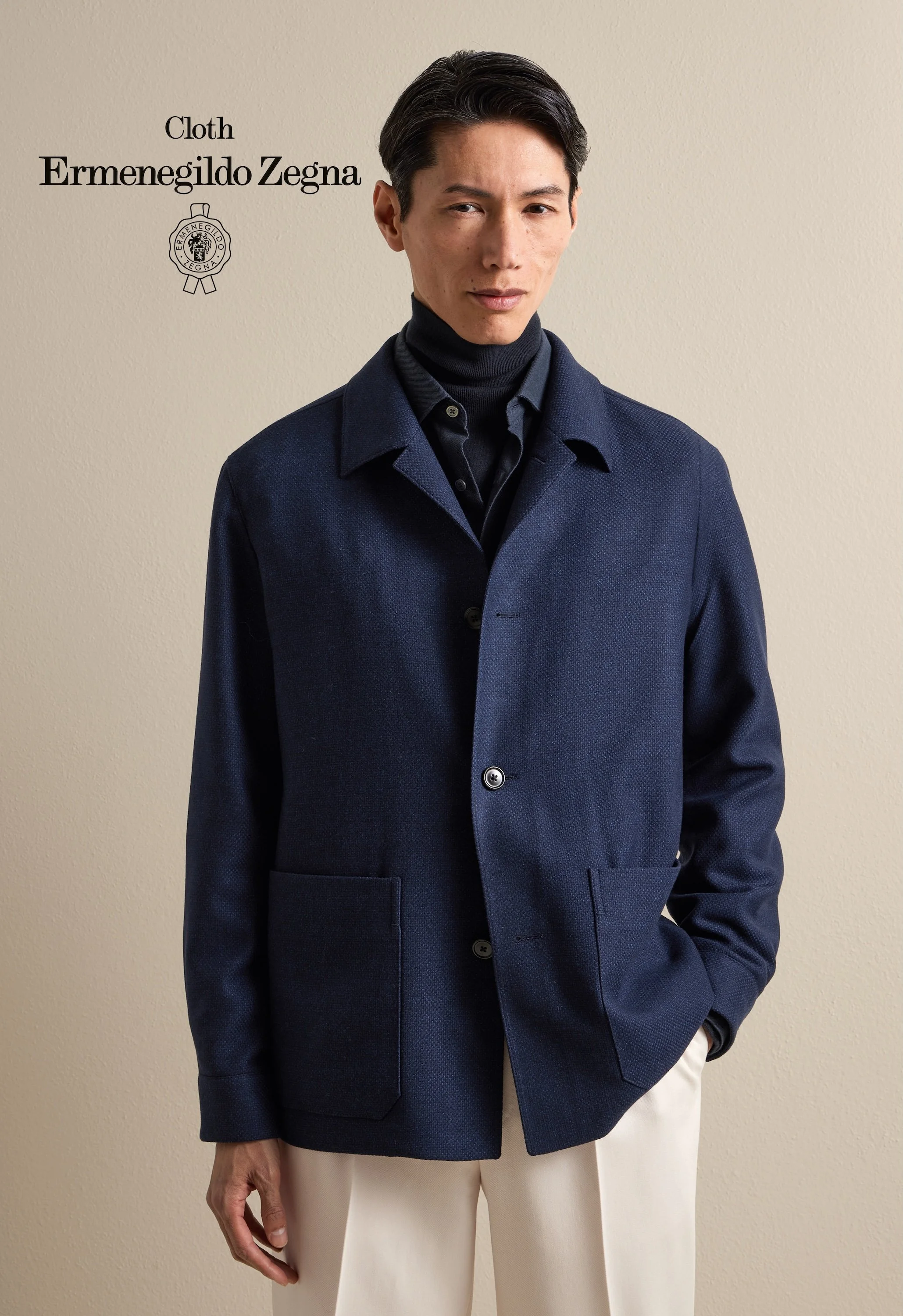 A man wearing a dark navy Zegna coat over a black turtleneck sweater, beige pants, standing against a plain beige wall.