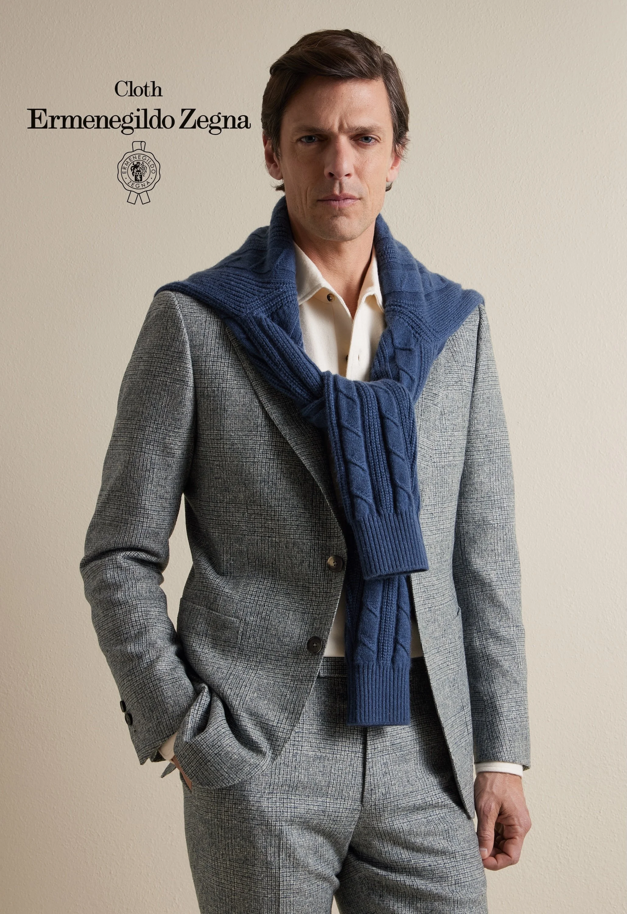 A man wearing a plaid gray suit with a beige shirt and a blue knit sweater draped around his shoulders, standing against a beige wall, with the words "Cloth Ermenegildo Zegna" and a logo on the left side.