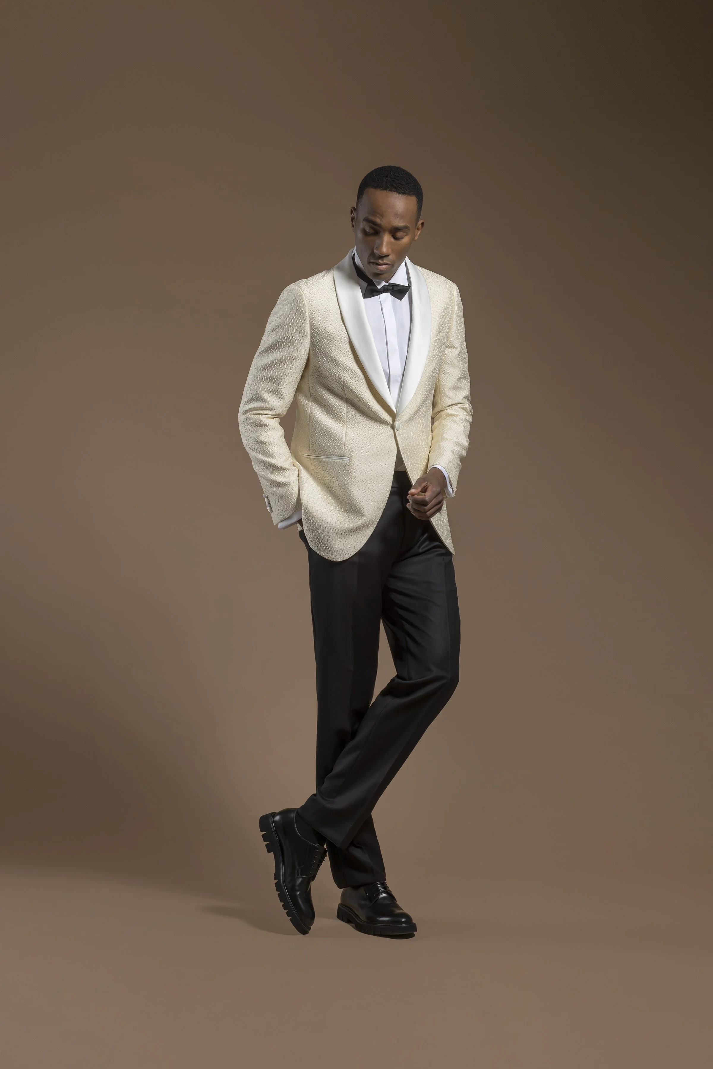 A man wearing a Scabal white tuxedo jacket with a black bow tie, black dress pants, and black shoes standing against a plain brown background.