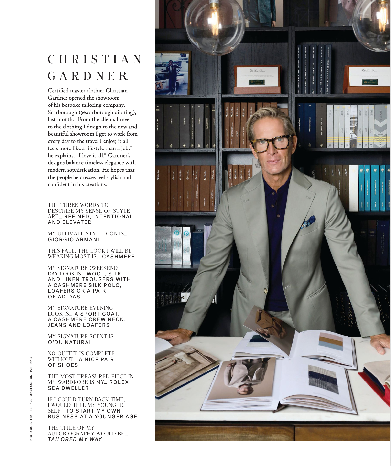 Christian Gardner standing in the Scarborough Custom Tailoring showroom in modern, stylish attire with glasses sitting at a table with fashion magazines open, surrounded by shelves with books and framed photos, and hanging decorative lights.