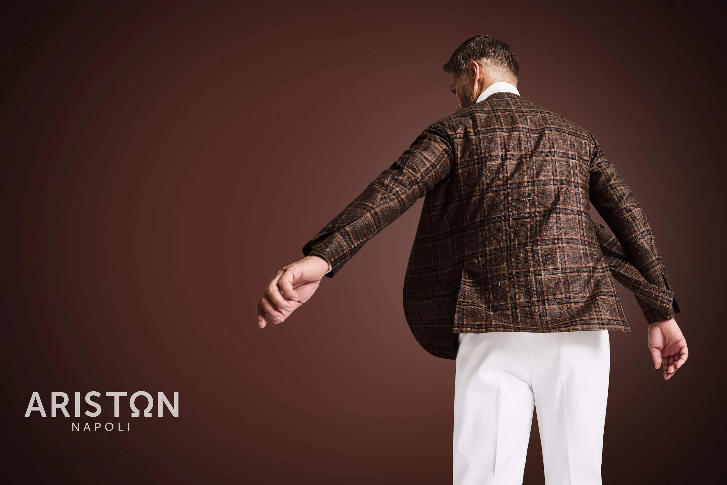 A man wearing an Ariston brown checkered blazer and white pants stands with his back to the camera against a brown background, slightly turning to the side with arms outstretched.
