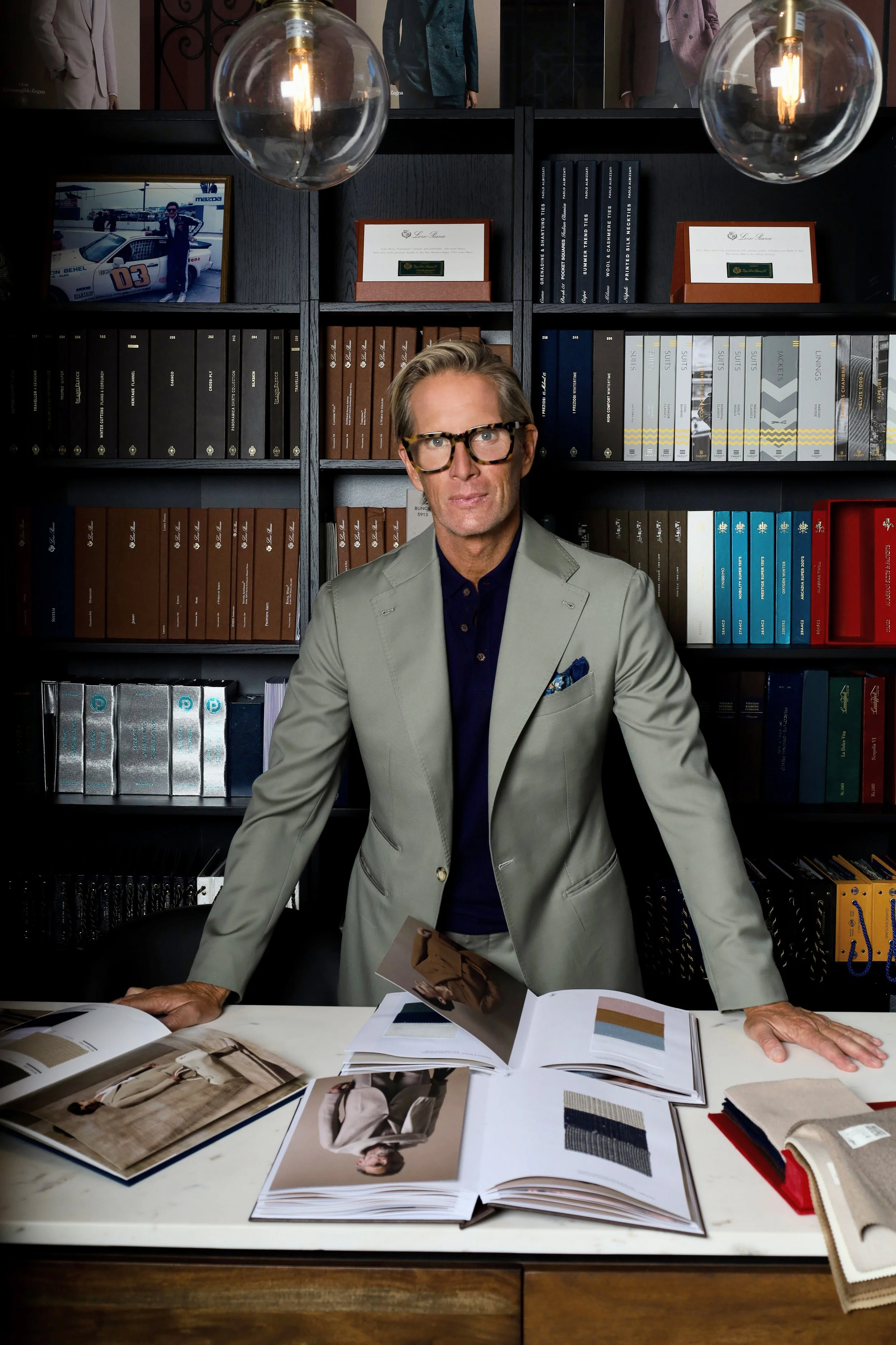 Christian Gardner standing in the Scarborough Custom Tailoring showroom wearing glasses and a light gray suit sits at a desk with fashion magazines and fabric swatches, in front of a bookshelf filled with books, awards, and framed photos.