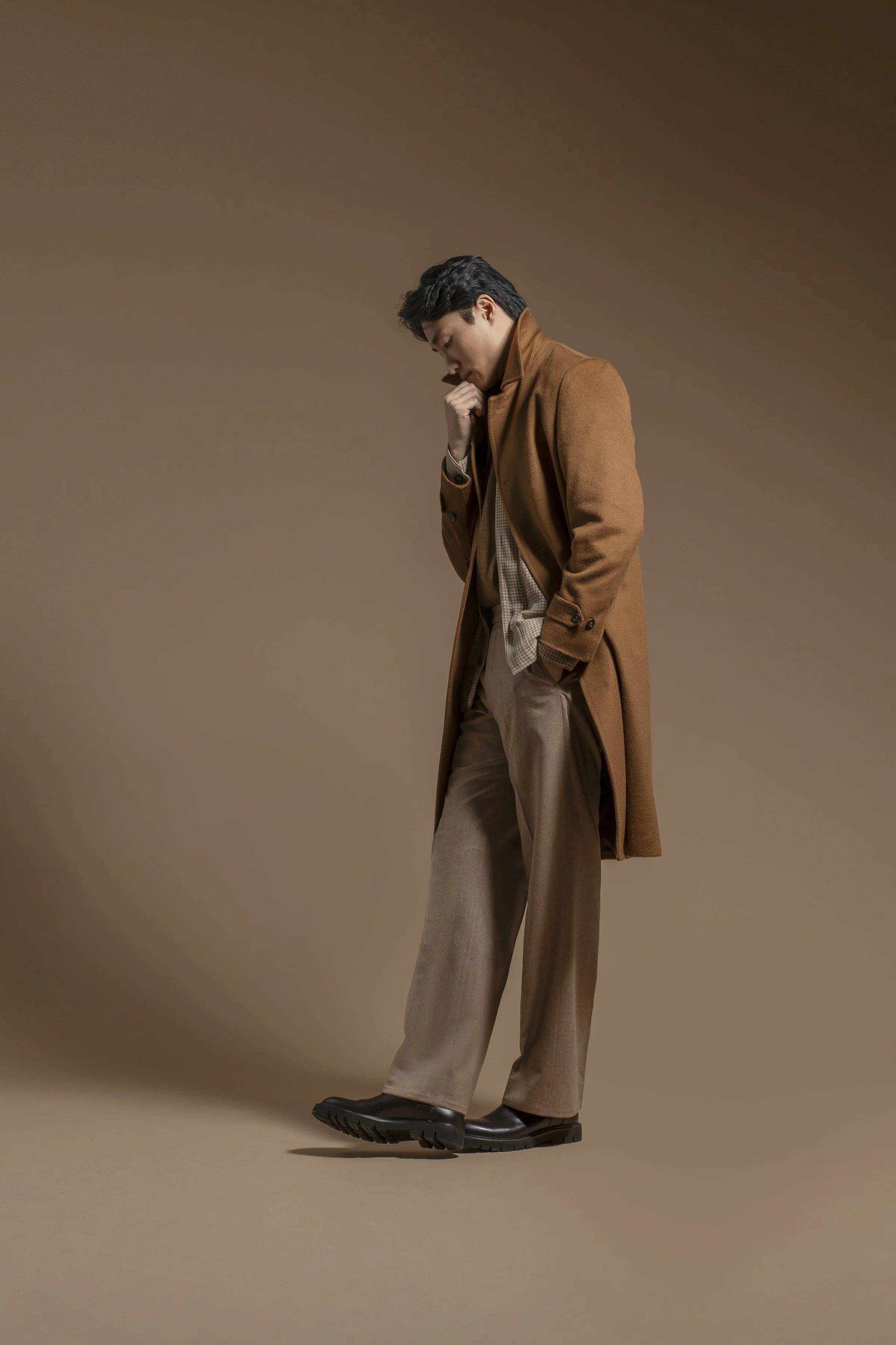 A young man poses against a plain, muted brown background. He is dressed in a stylish tan Scabal overcoat, beige trousers, and dark shoes. He is standing with his head slightly bowed and his right hand resting on his chin.