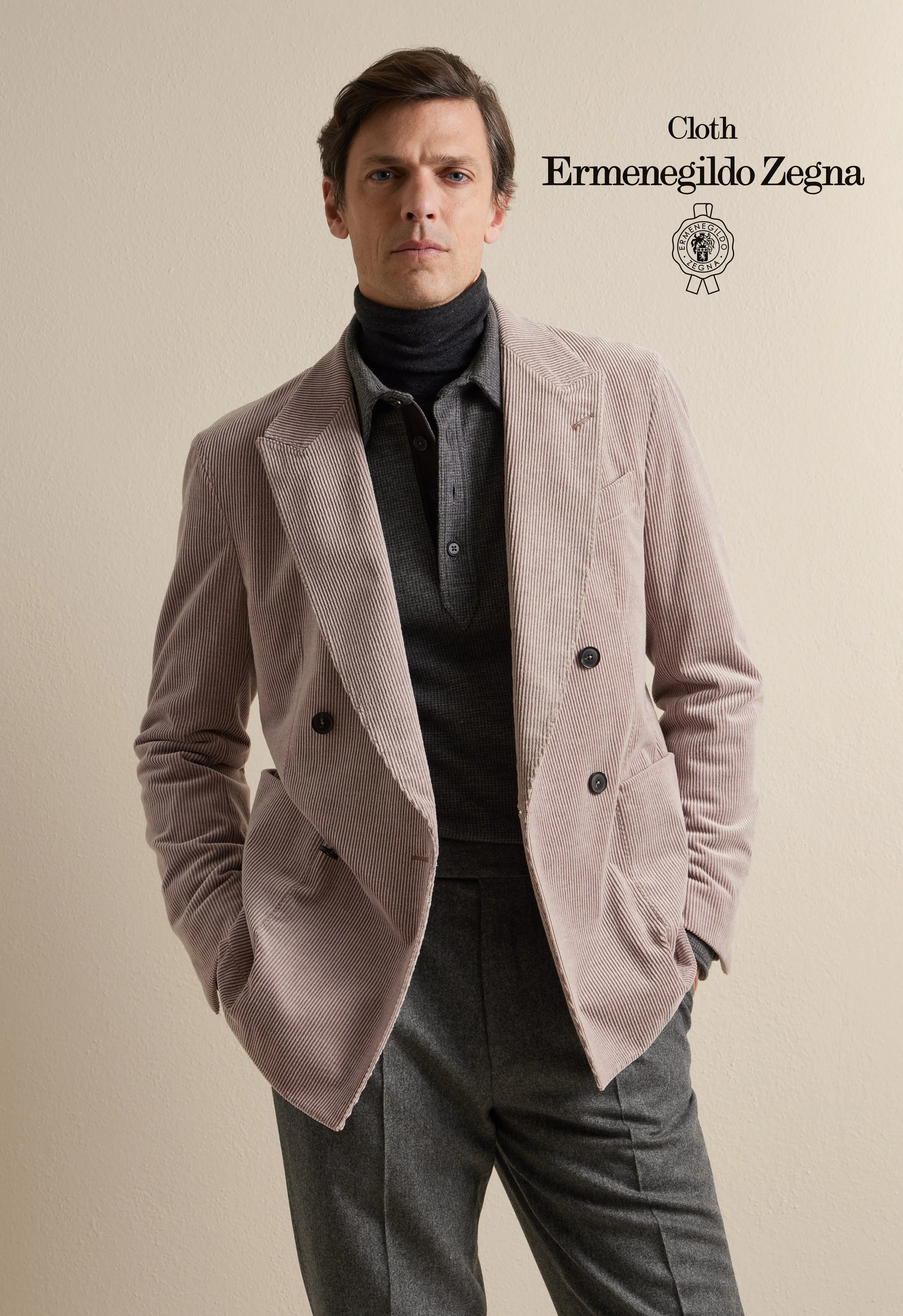 A man in a Zegna light pink double-breasted blazer, black turtleneck, and gray trousers standing against a beige wall.