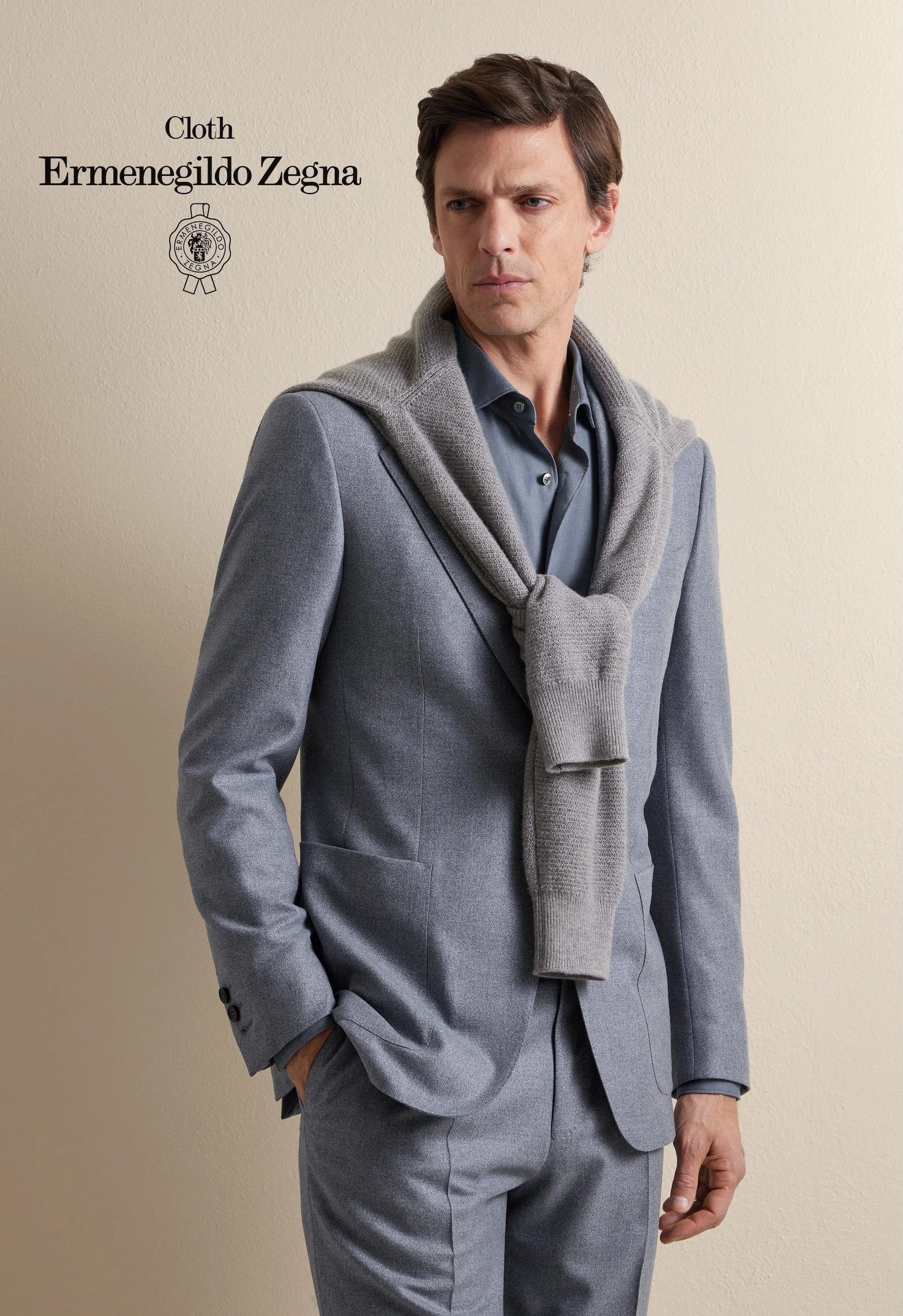 A man in a tailored grey Zegna suit with a grey sweater draped over his shoulders, standing against a plain beige wall.