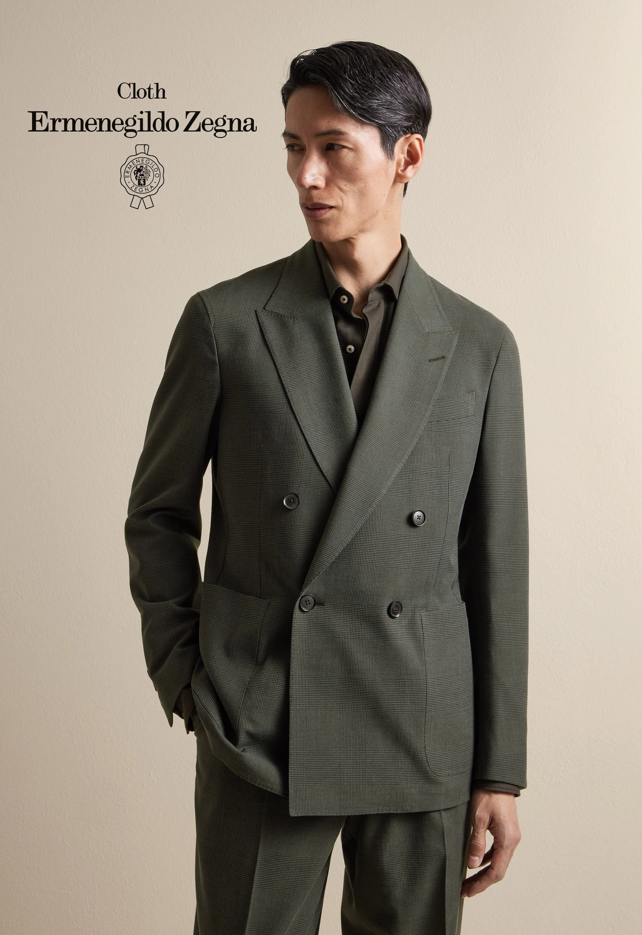 A man wearing a green plaid double-breasted Zegna suit with a black shirt, standing against a plain beige wall.