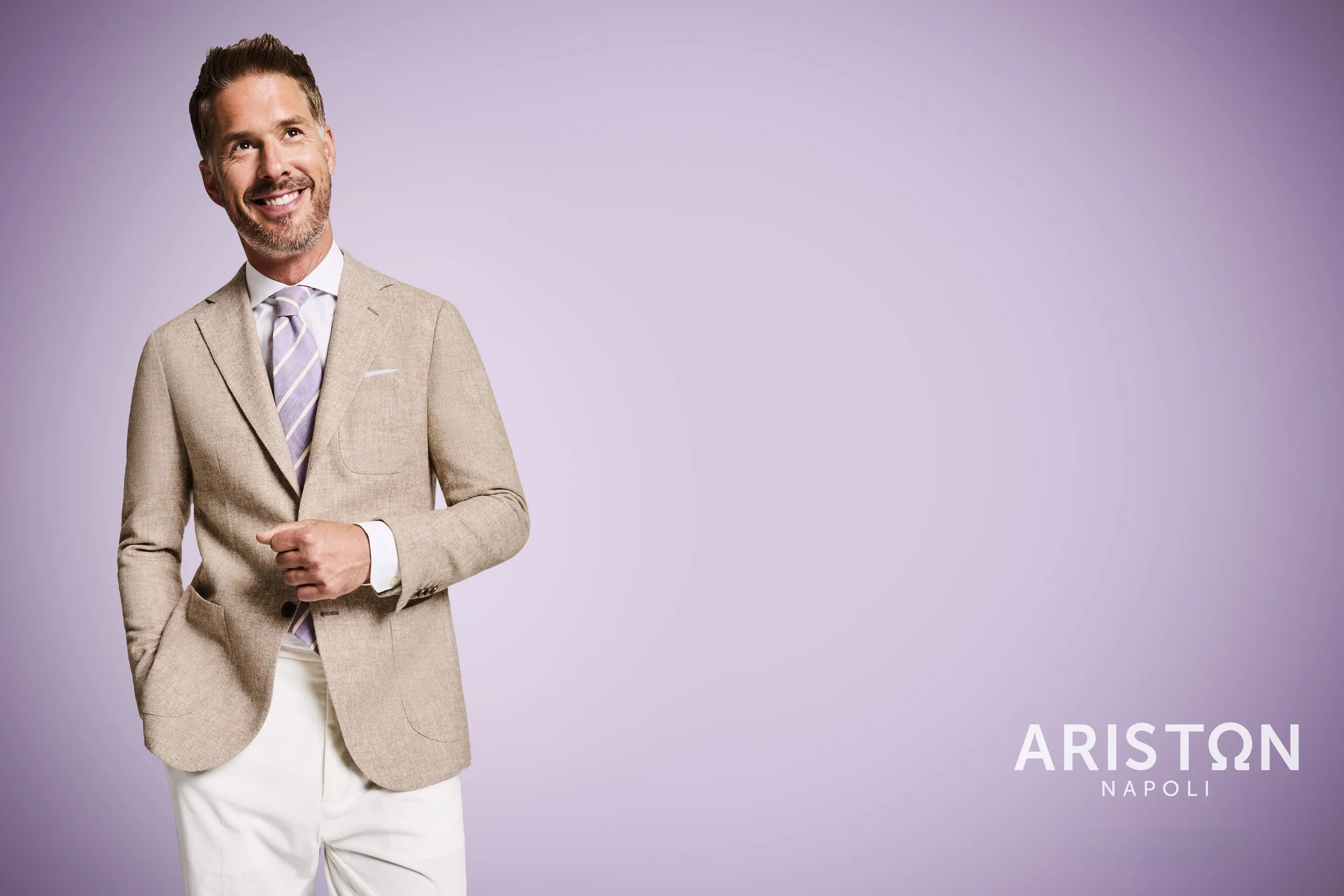 A man with brown hair and a beard wearing a beige suit jacket with a white shirt and a lavender striped tie, standing against a light purple background. The text ''ARISTON NAPOLI'' is visible at the bottom right corner.
