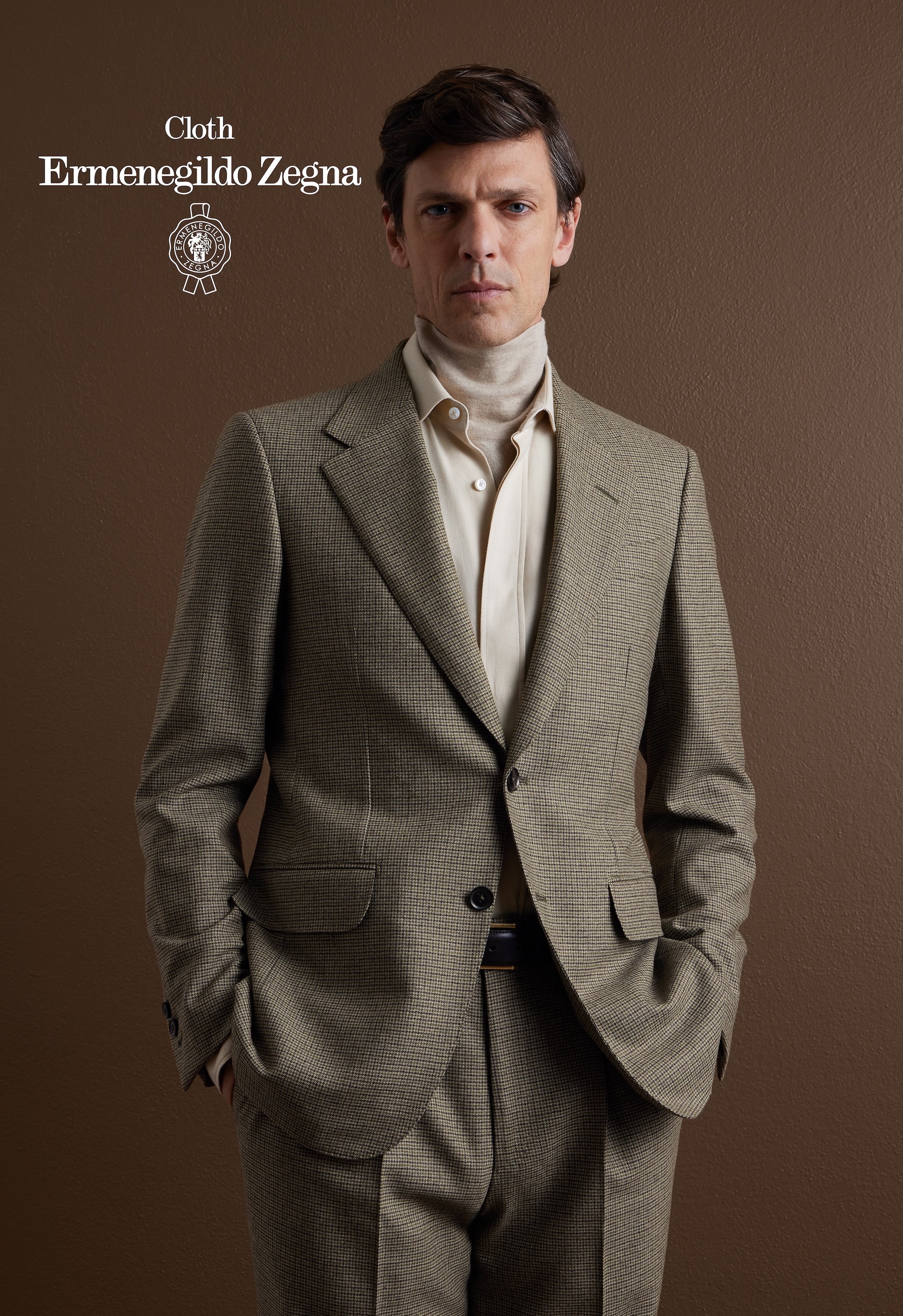 A man in a beige plaid Zegna suit with a turtleneck shirt stands against a brown wall, with one hand in his pocket and serious expression.