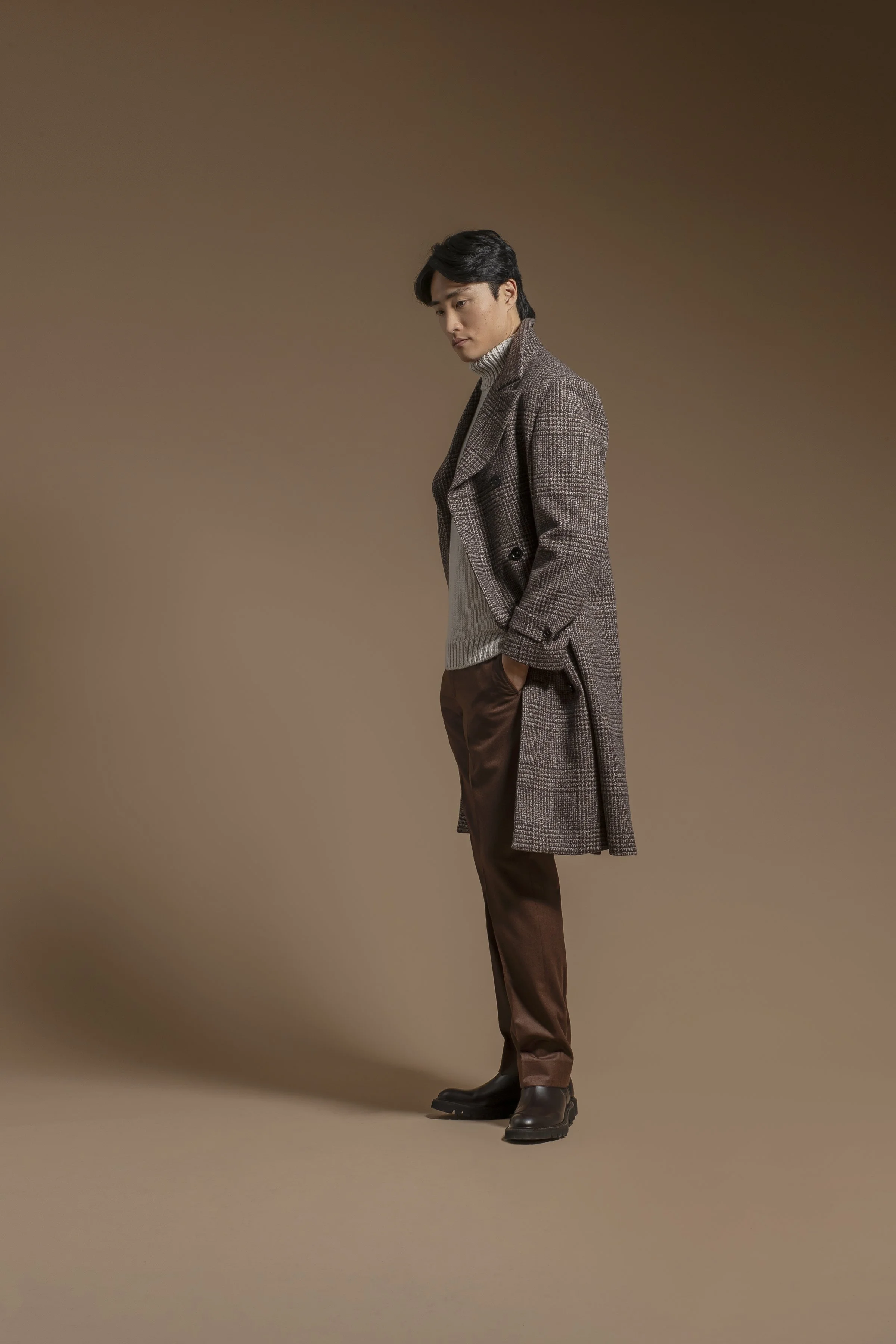 A man standing in a brown studio background, dressed in a long dark plaid Scabal overcoat, a beige turtleneck sweater, brown pants, and black leather boots.