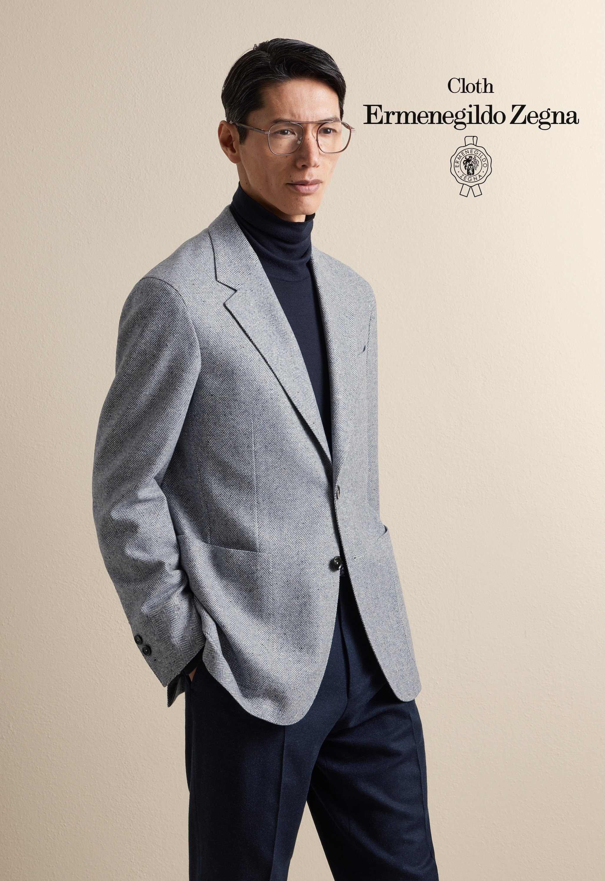 A man wearing glasses, a black Zegna turtleneck, and a tailored light gray blazer with dark trousers, standing against a beige wall.