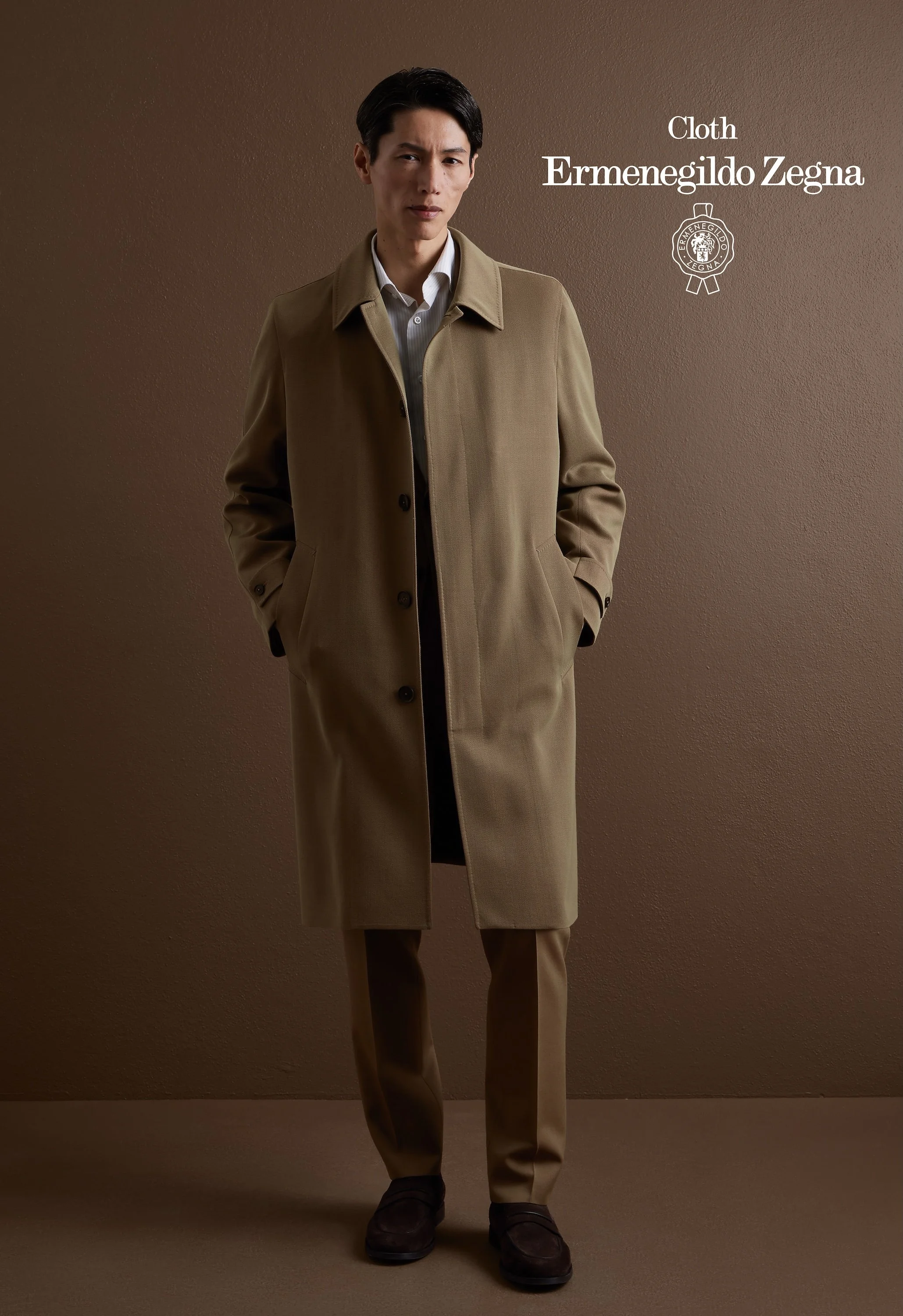 A man standing against a brown wall wearing a tan overcoat, tan pants, dark shoes, and a white shirt. Text on the right reads "Cloth Ermenegildo Zegna" with a logo below.