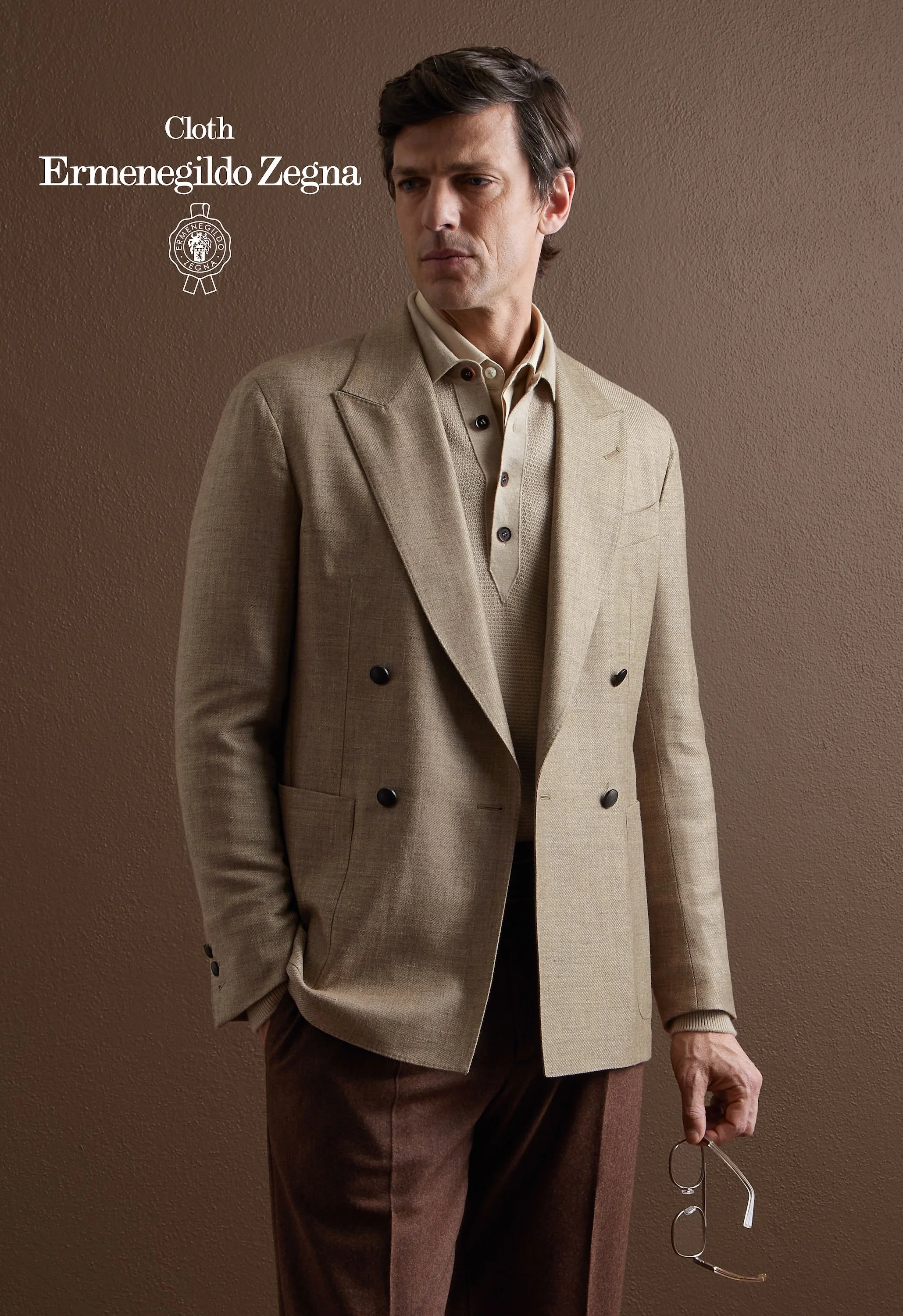 A man dressed in a beige Zegna suit jacket, a light-colored shirt, and brown pants, standing against a brown textured wall, holding glasses in his right hand.