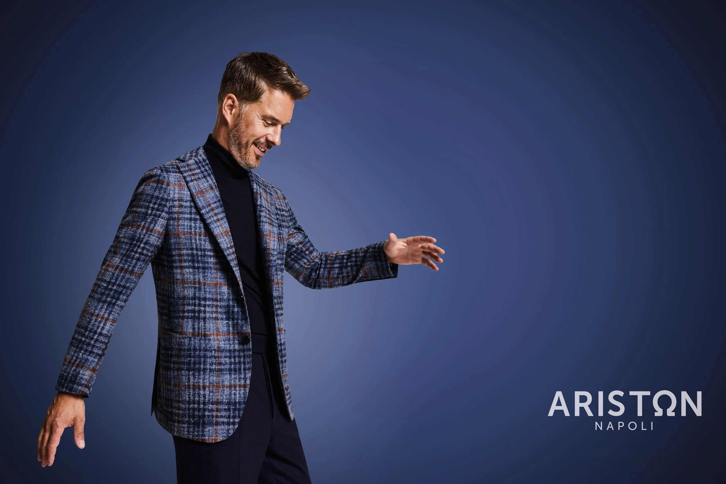 A man with brown hair and a beard wearing a black turtleneck and a plaid blazer, smiling and looking downward in front of a dark blue background. The text in the bottom right corner reads 'ARISTON NAPOLI'.