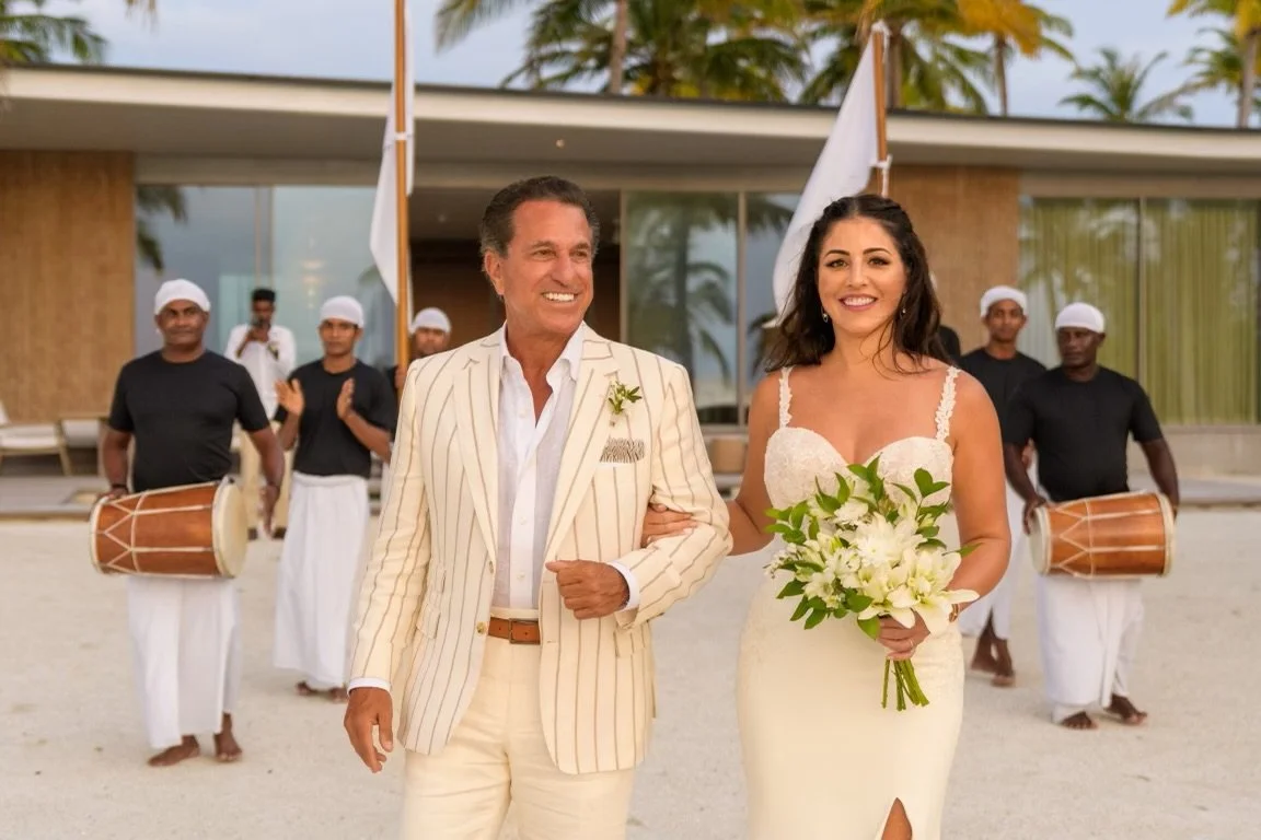 A bride in a white wedding dress holding a bouquet of white flowers walking arm-in-arm with a man in a light-colored striped Scarborough Custom Tailoring suit, smiling at a beachside wedding, with drummers and servers in the background.