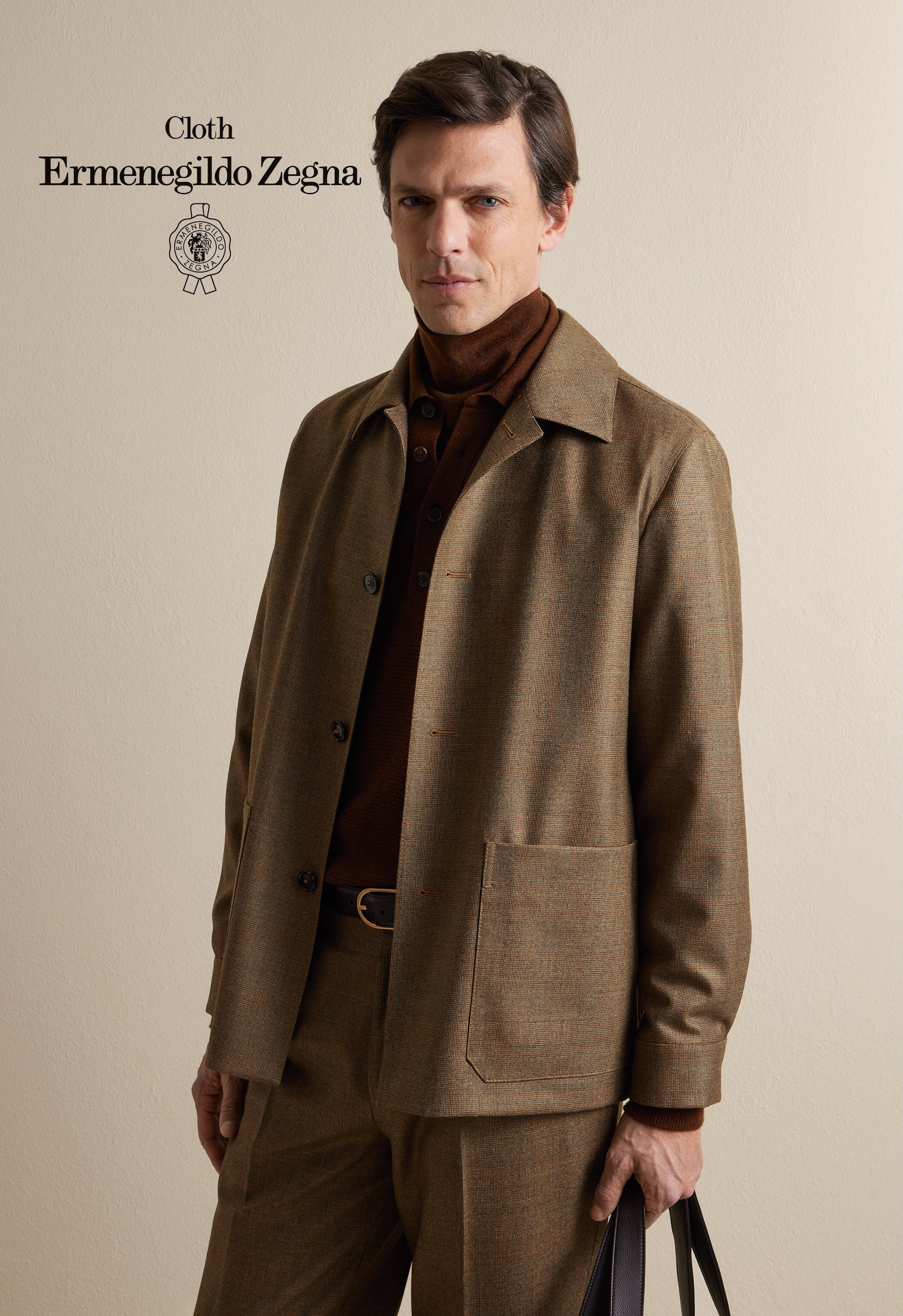 A man dressed in brown plaid suit, holding black leather bag, standing against plain beige wall with text 'Cloth Ermenegildo Zegna' and logo.