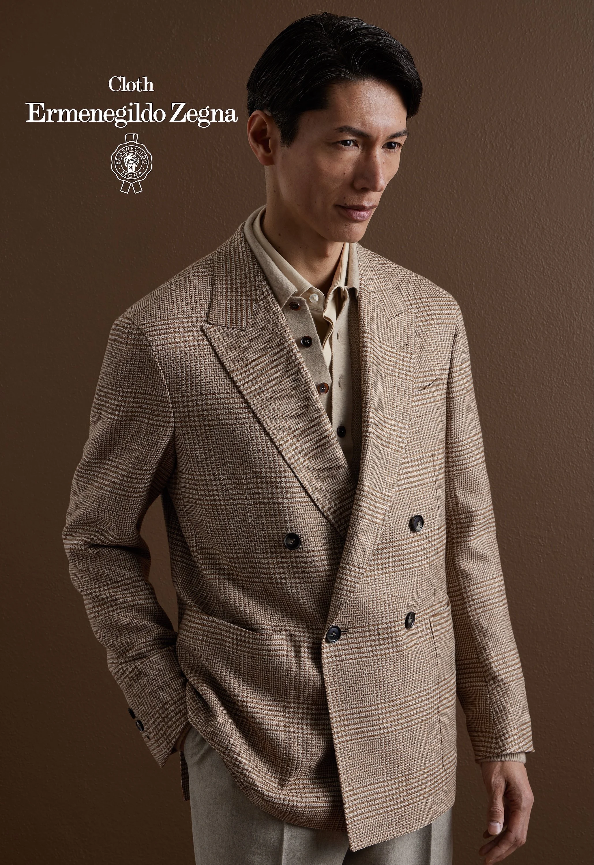 A man wearing a beige plaid double-breasted Zegna blazer and a light beige shirt, standing against a brown wall.