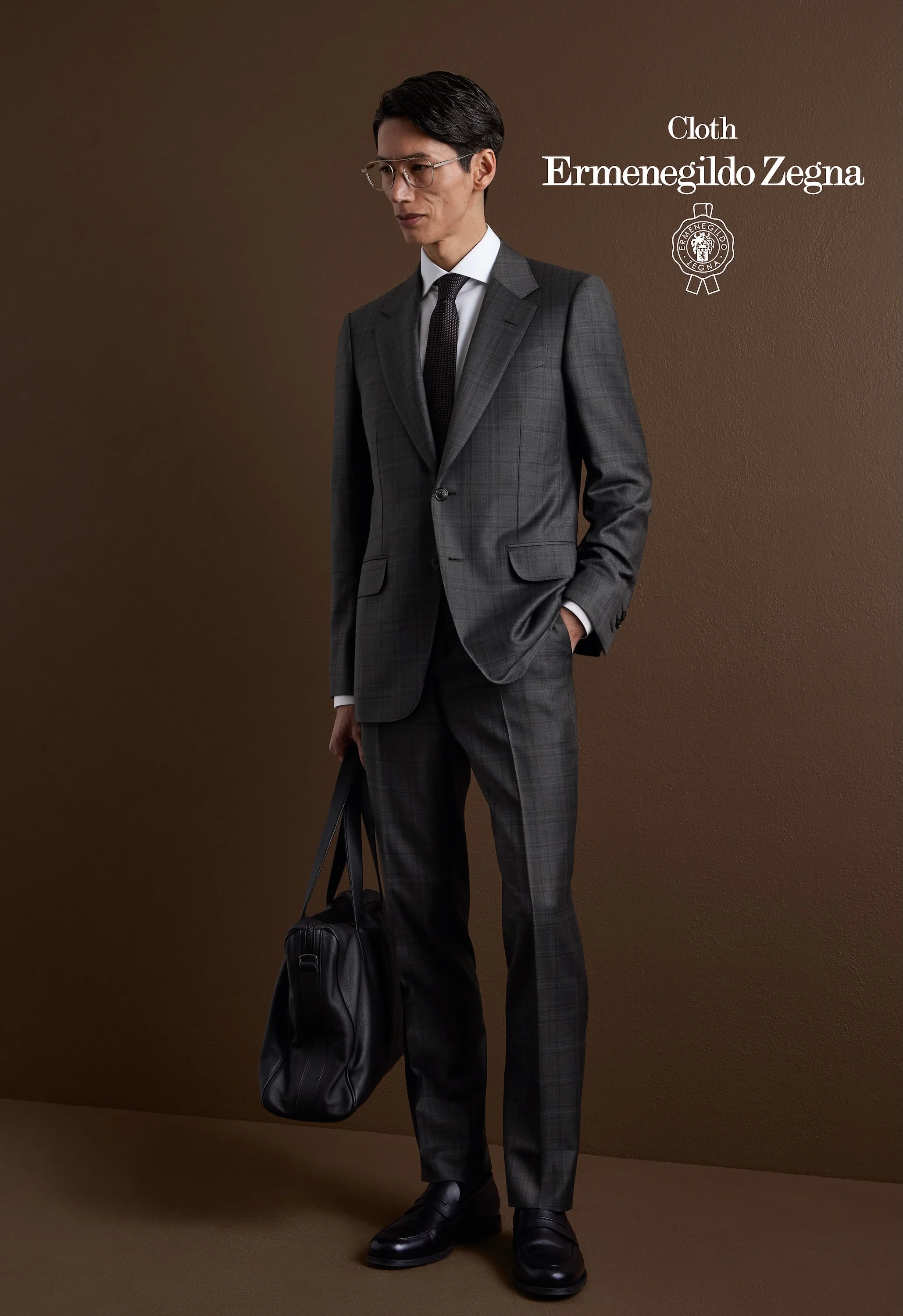 A man dressed in a grey plaid Zegna suit with a white shirt and black tie, wearing glasses, standing against a brown background with a black bag on the floor and holding a black briefcase.