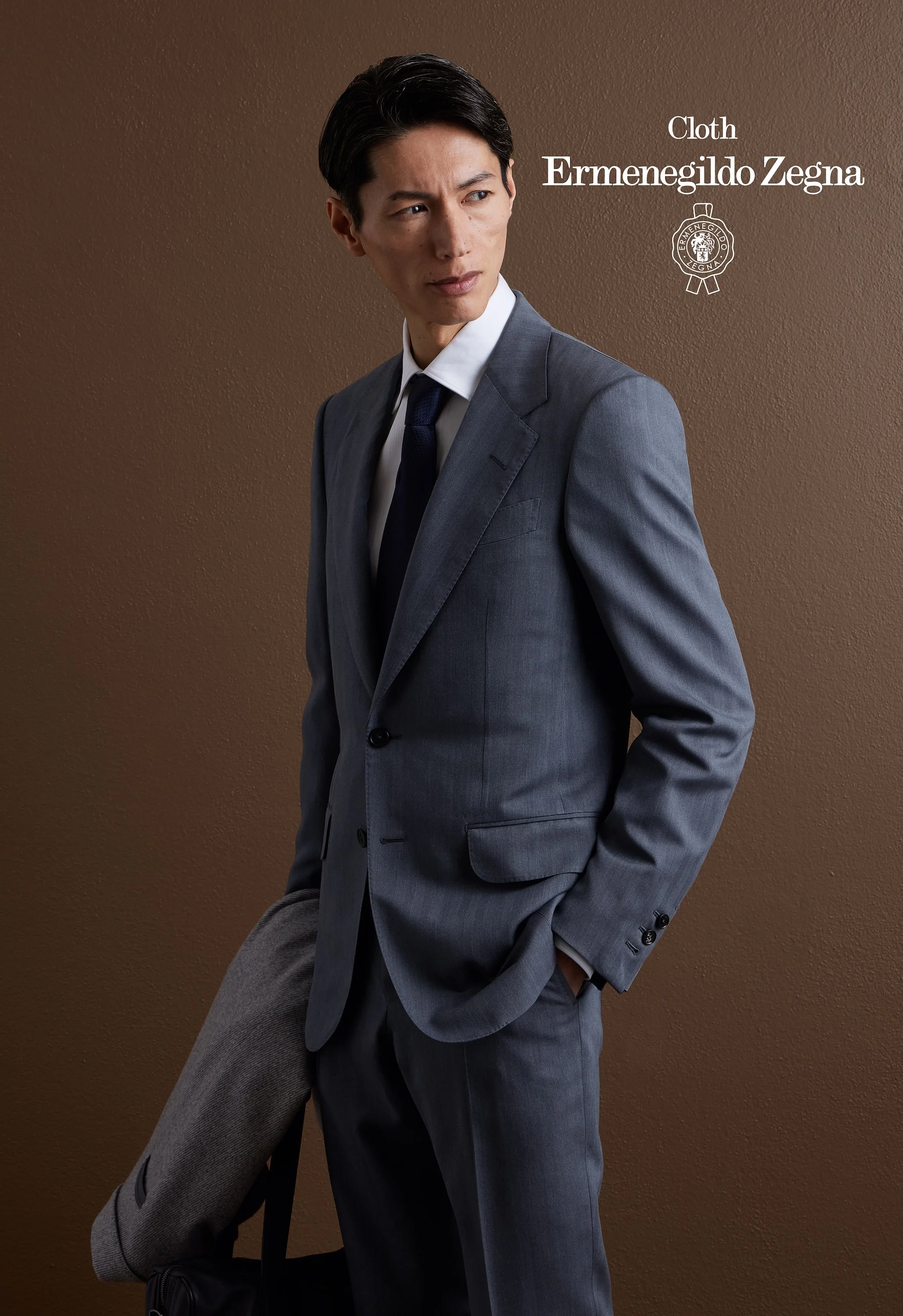 A man in a gray suit with a white shirt and navy tie, standing against a brown textured background, holding a black bag with his left hand in his pocket, looking to his left, with text on the right side that reads 'Cloth Ermenegildo Zegna' and a logo
