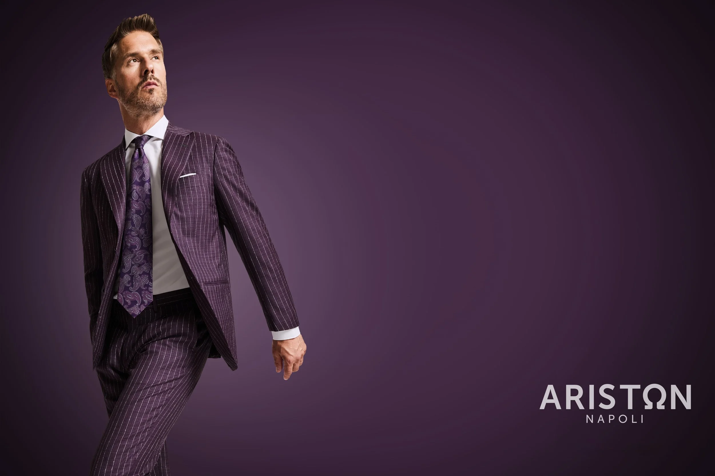 A man wearing a purple pinstripe suit and a purple patterned tie standing against a purple background. The text on the bottom right reads "Ariston Napoli."