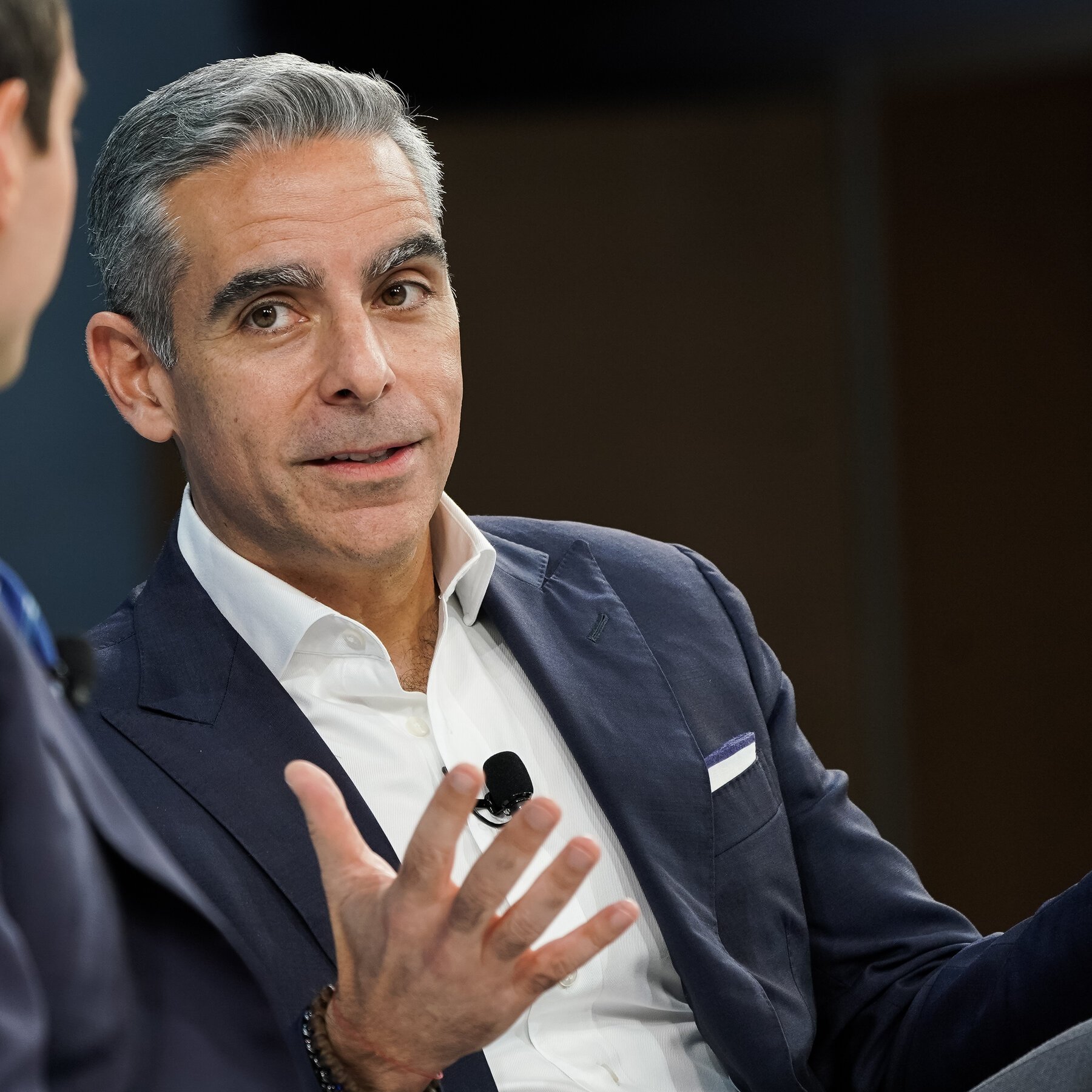 A middle-aged man with gray hair wearing a navy-blue suit and a white collared shirt, engaged in conversation during a formal meeting or interview.