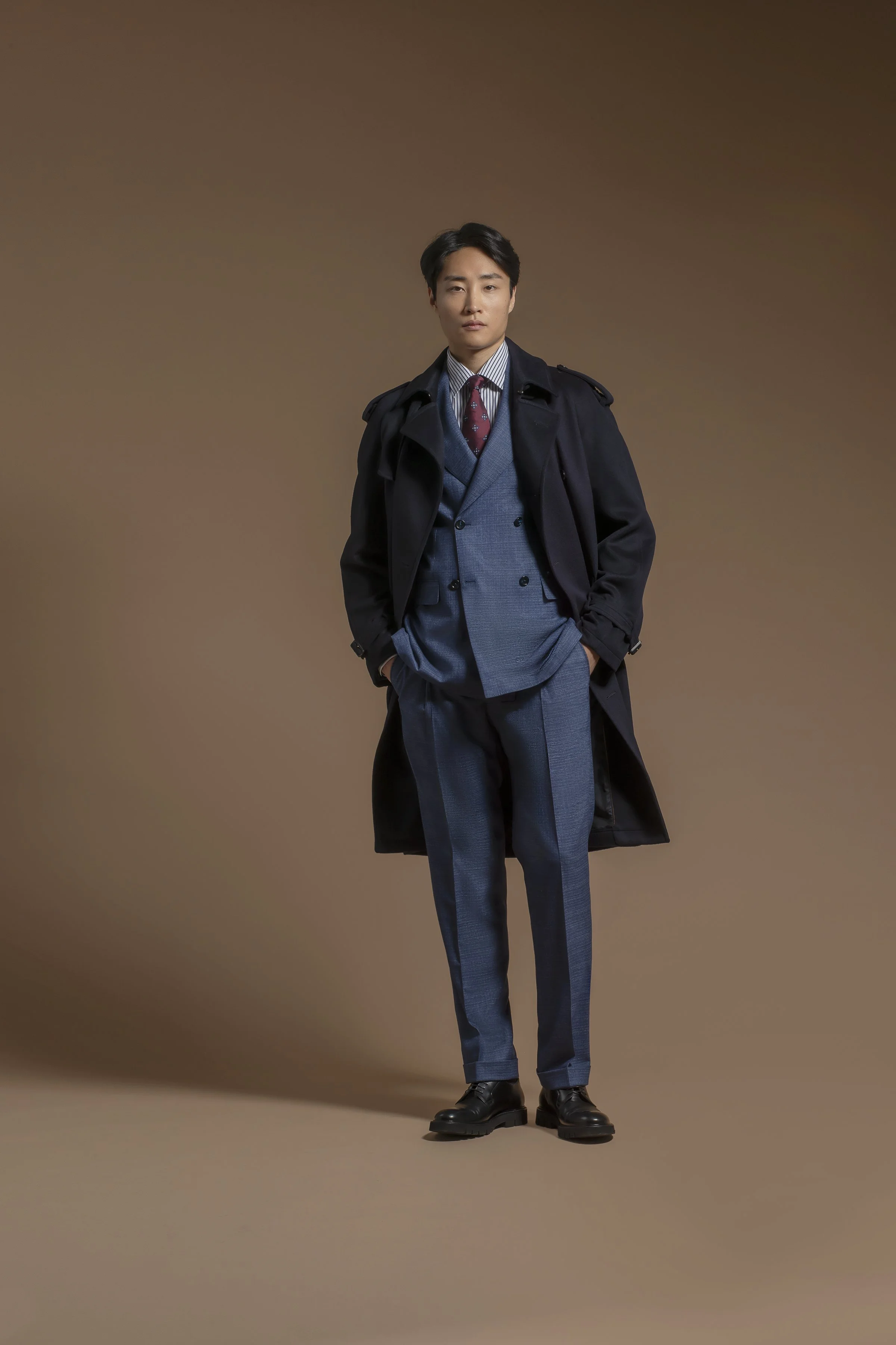 A young man standing against a plain brown background, wearing a dark Scabal trench coat, a blue blazer, a white shirt, a red patterned tie, and dress shoes, with his left hand in his pocket.