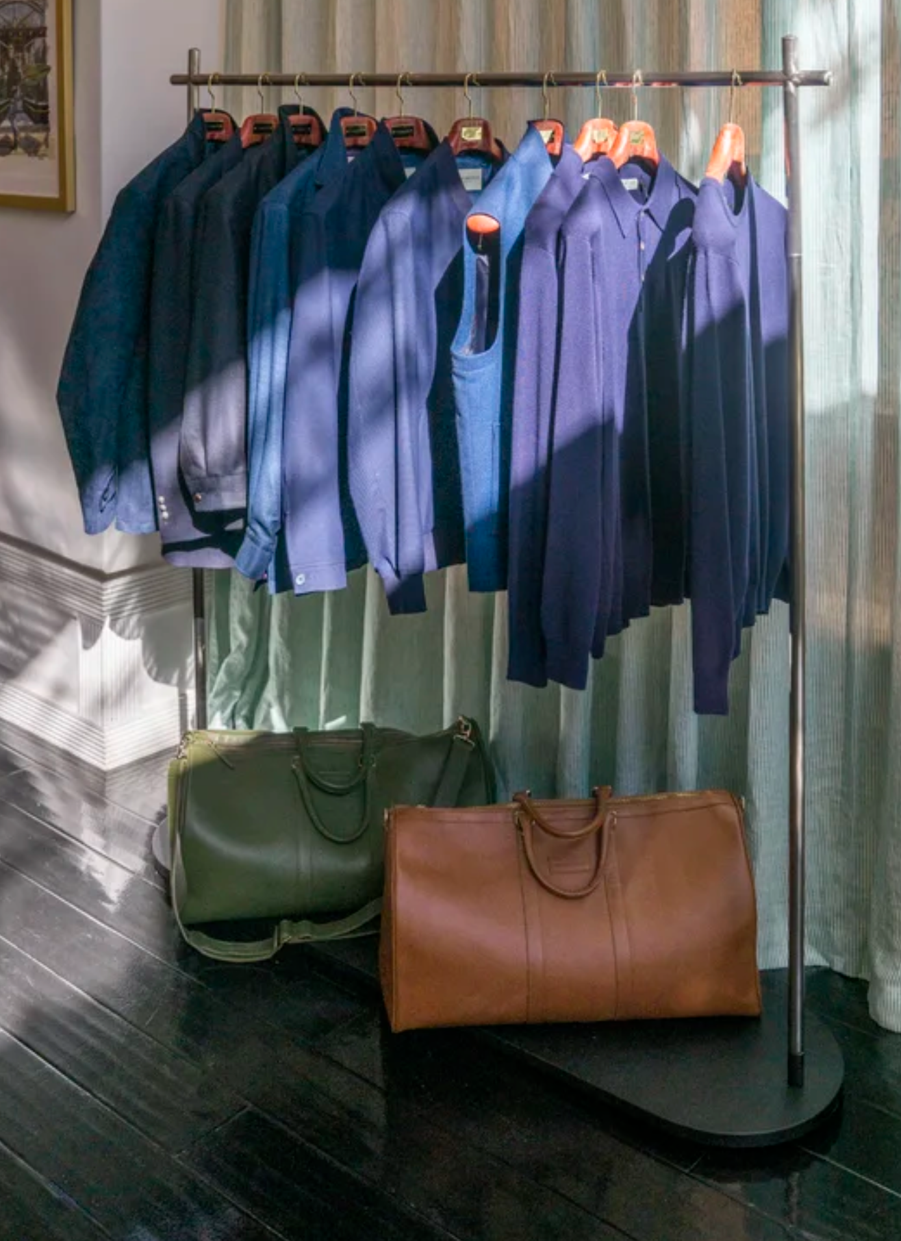 A clothing rack with several blue and navy shirts hanging on hangers, and two large custom leather bags, one green and one brown, placed on the dark wooden floor below the rack in the Scarborough showroom in San Francisco..