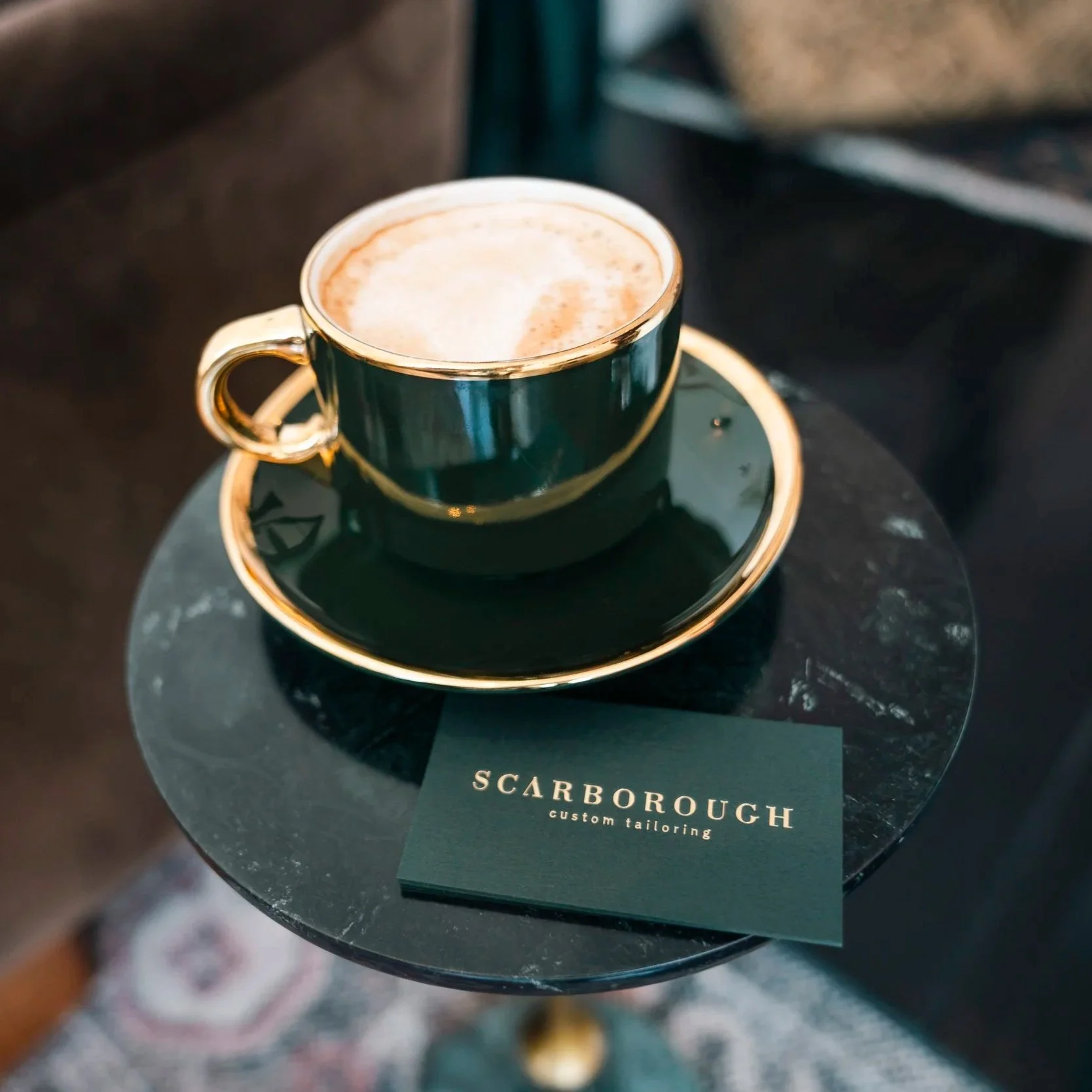 Dark green cup with gold handle and rim filled with frothy cappuccino, placed on a black saucer on a marble table, with a black card from Scarborough Custom Tailoring underneath.