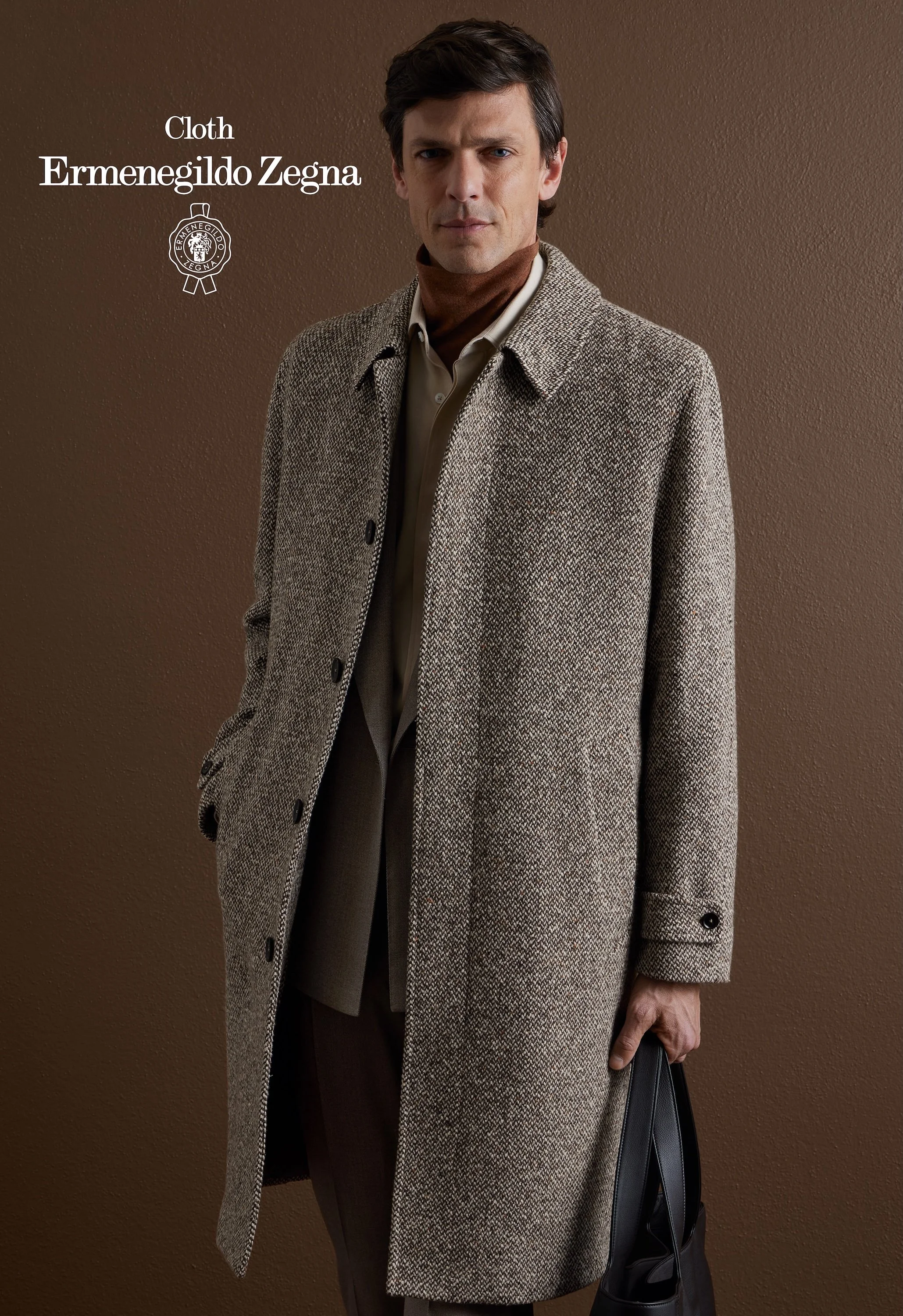 A man in a beige Zegna coat holding a black bag, standing against a brown wall.