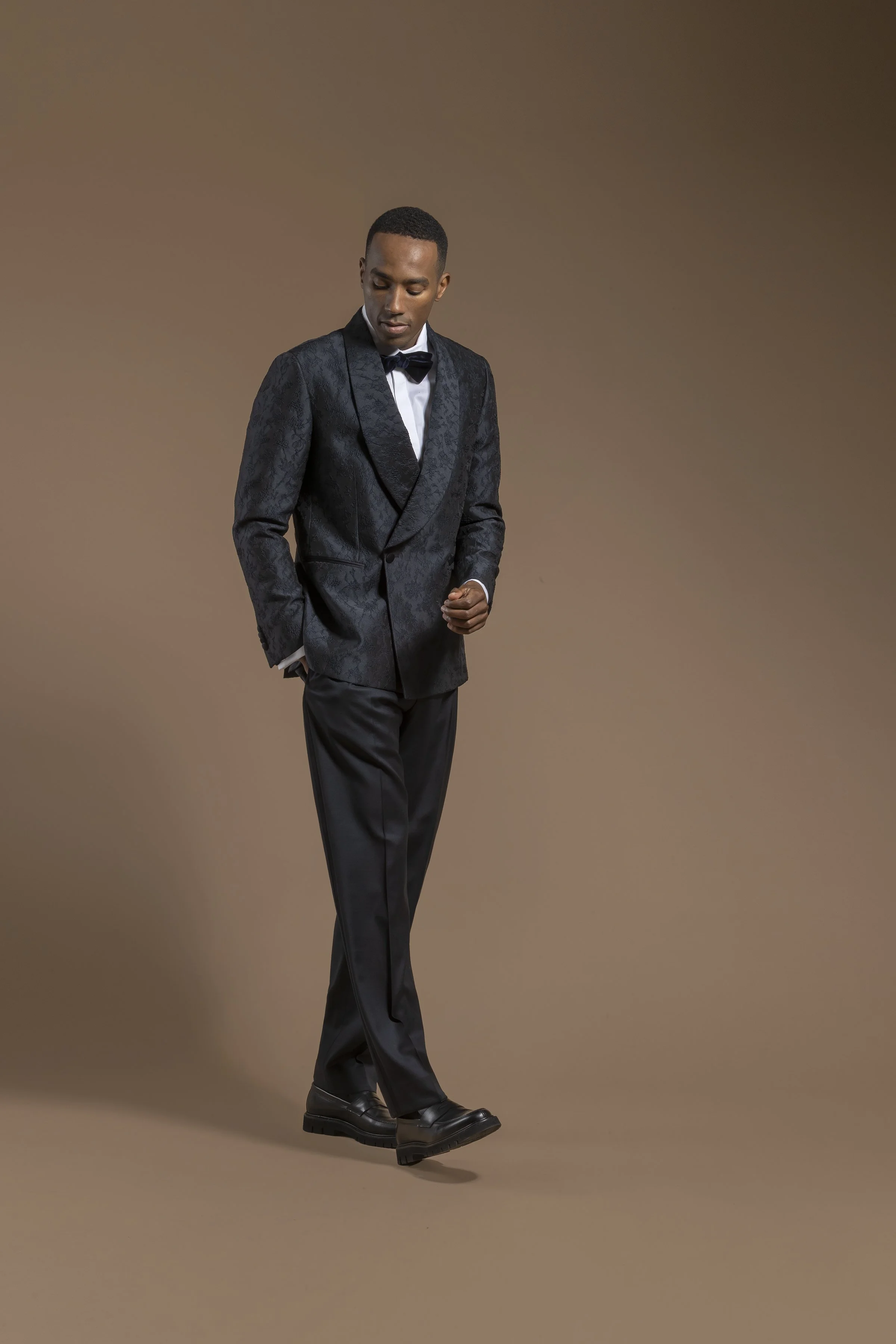 A man dressed in a Scabal black tuxedo with a patterned blazer, white shirt, and black bow tie, standing against a plain brown background, looking down with one hand in his pocket.