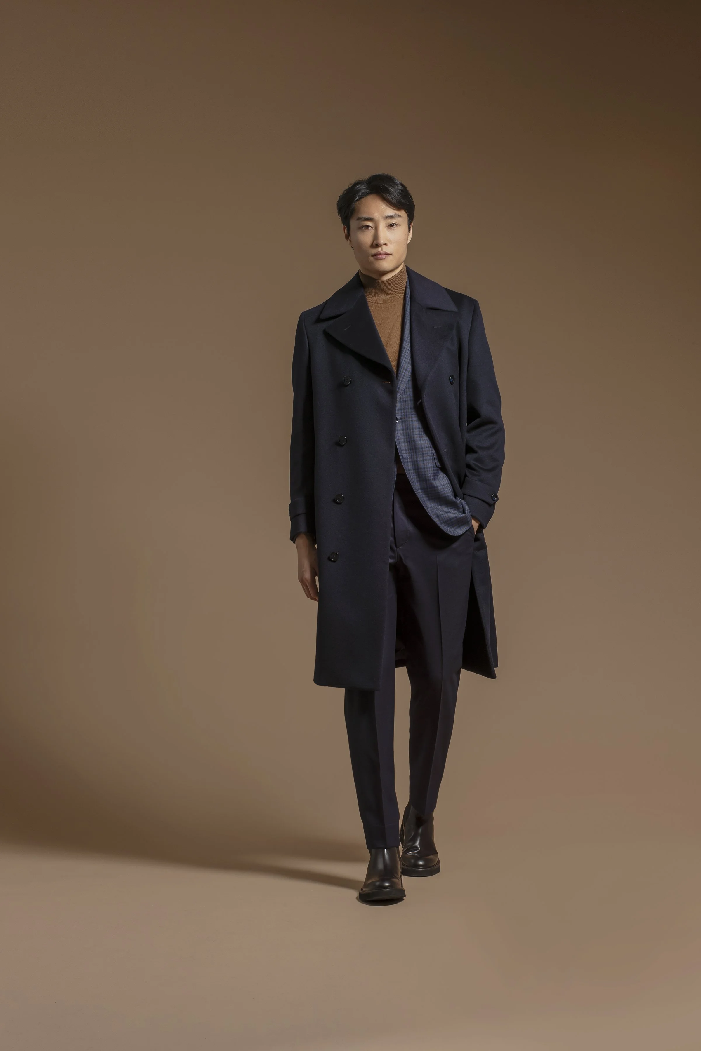 A young man with black hair, dressed in a brown Scabal turtleneck, navy blue trench coat, gray plaid scarf, navy trousers, and black leather boots, standing against a plain brown background.