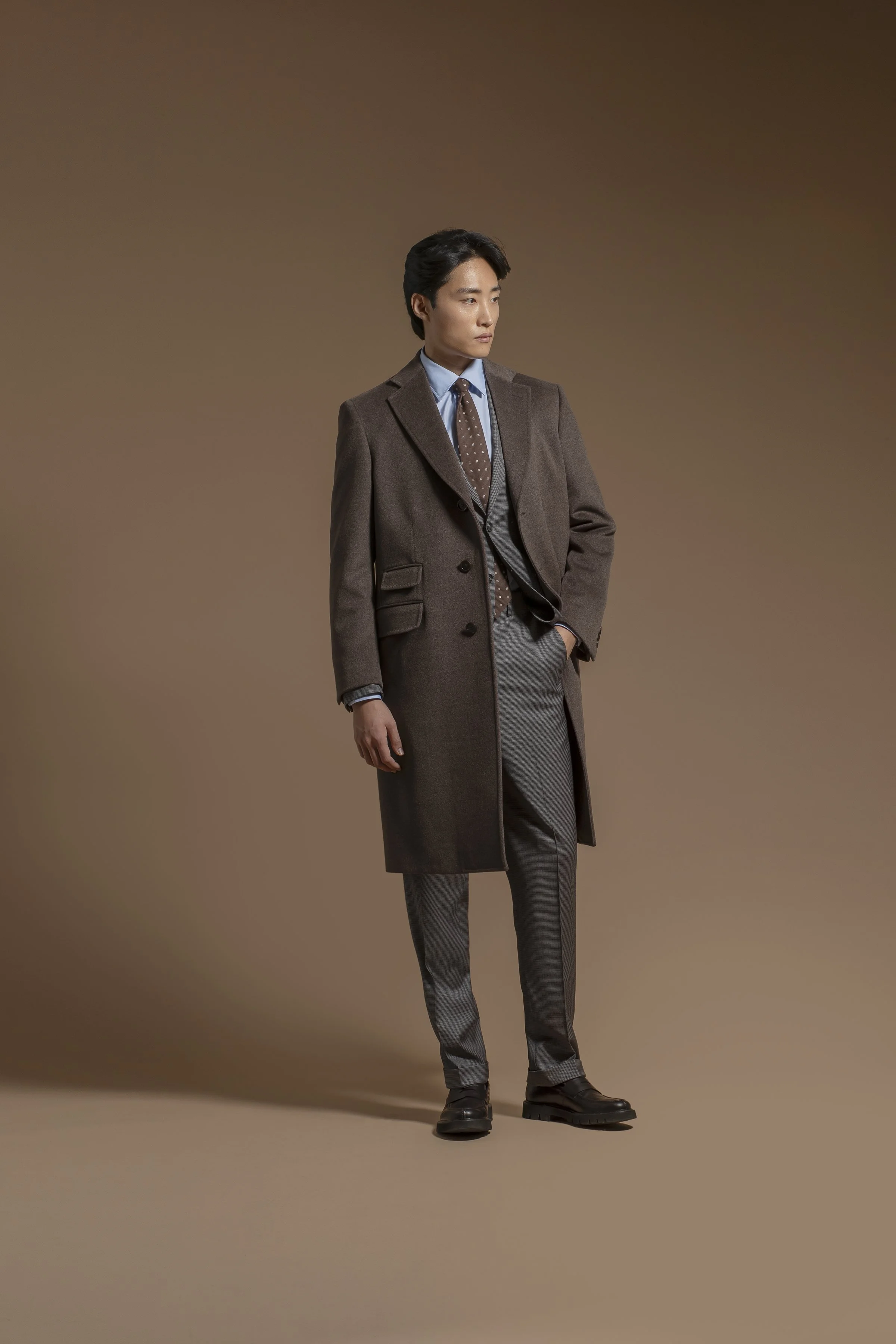 A young man with black hair styled to the side, wearing a brown Scabal overcoat, light blue shirt, and dark gray pants, standing against a plain brown background. He has one hand in his pocket and is looking off to the side.