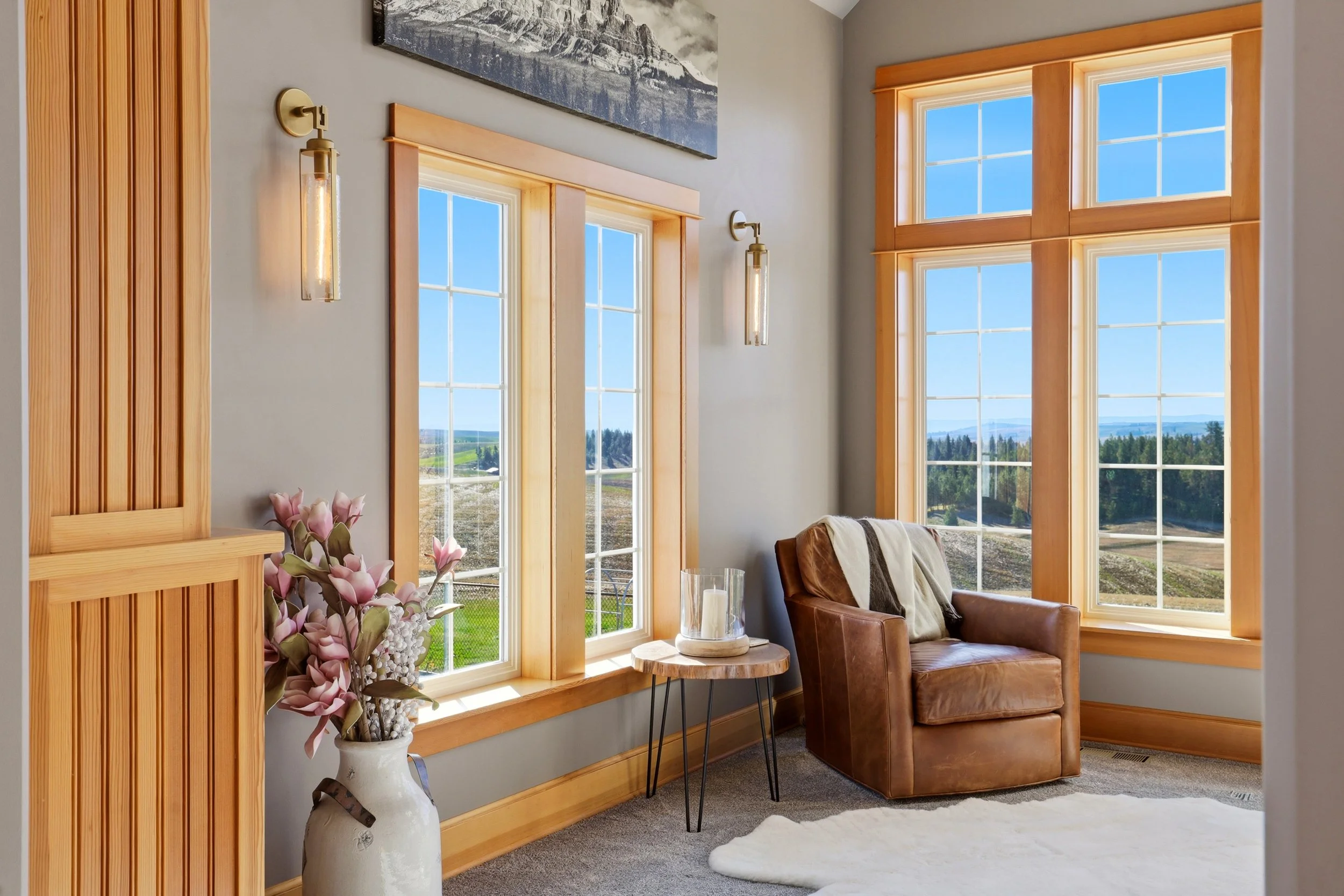A cozy living room with large windows showing a scenic outdoor landscape. The room features wooden trim around the windows, a brown leather armchair with a blanket, a small side table with candles, a vase of pink flowers, and a framed mountain landsc