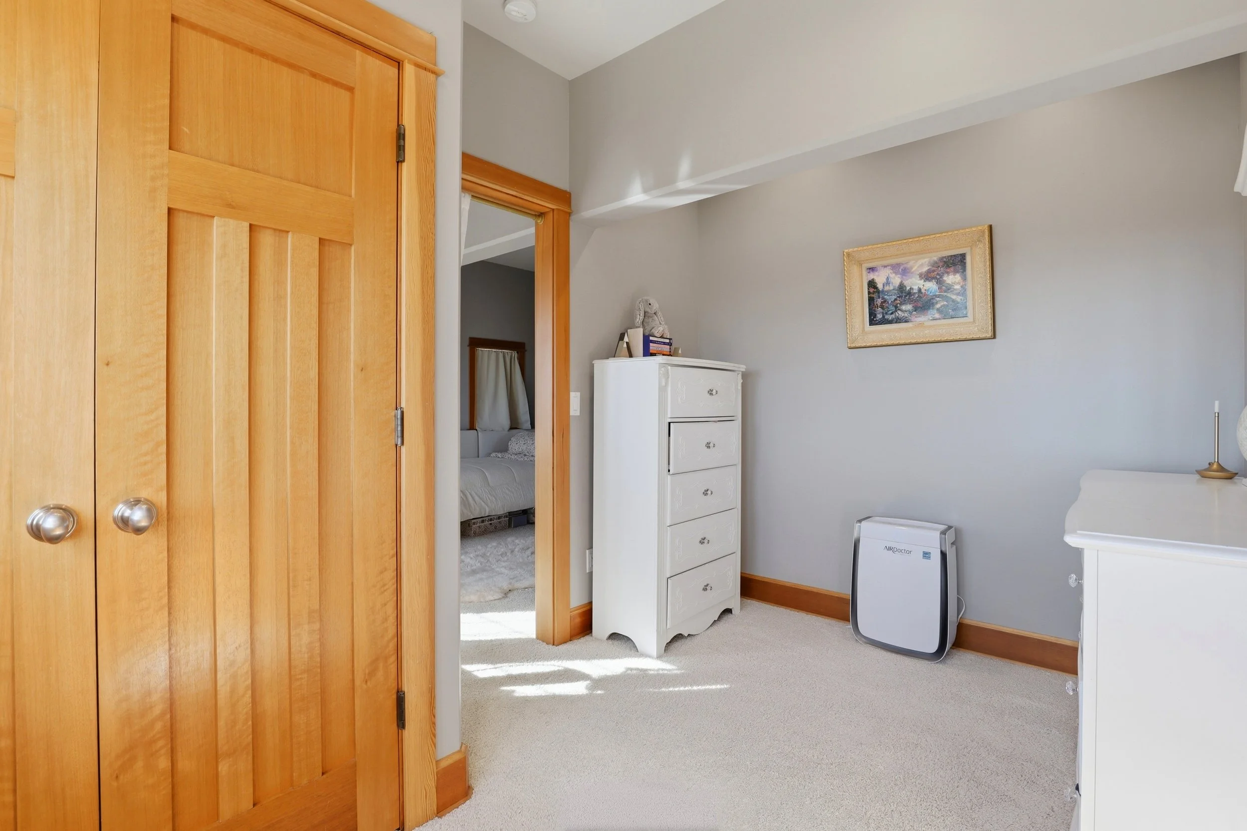 A view of a bedroom door open to a room with light gray walls, a white dresser, a framed painting, and a portable air purifier.