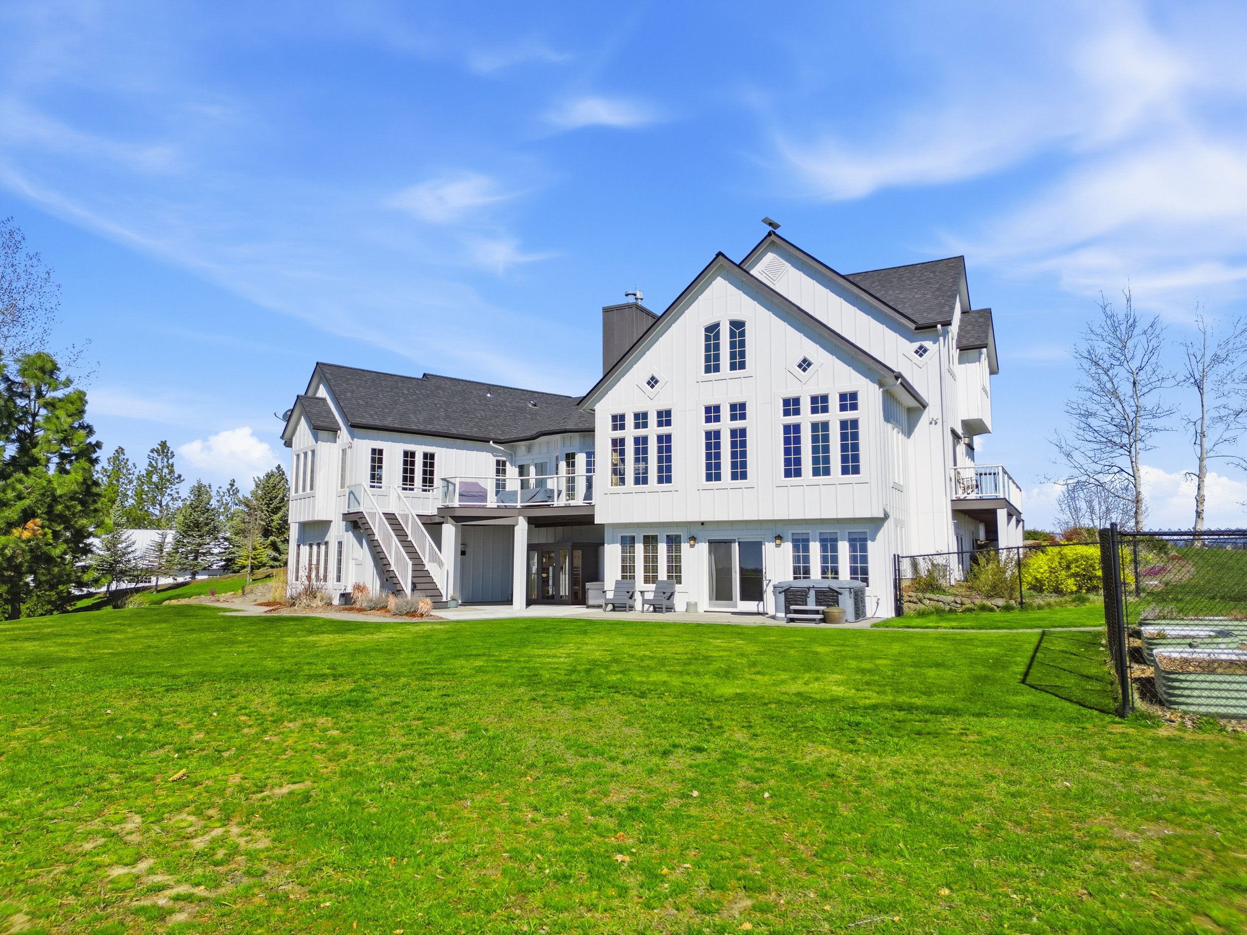 A large white house with multiple stories, many windows, and a dark gray roof, surrounded by a green lawn and trees under a blue sky with some clouds.