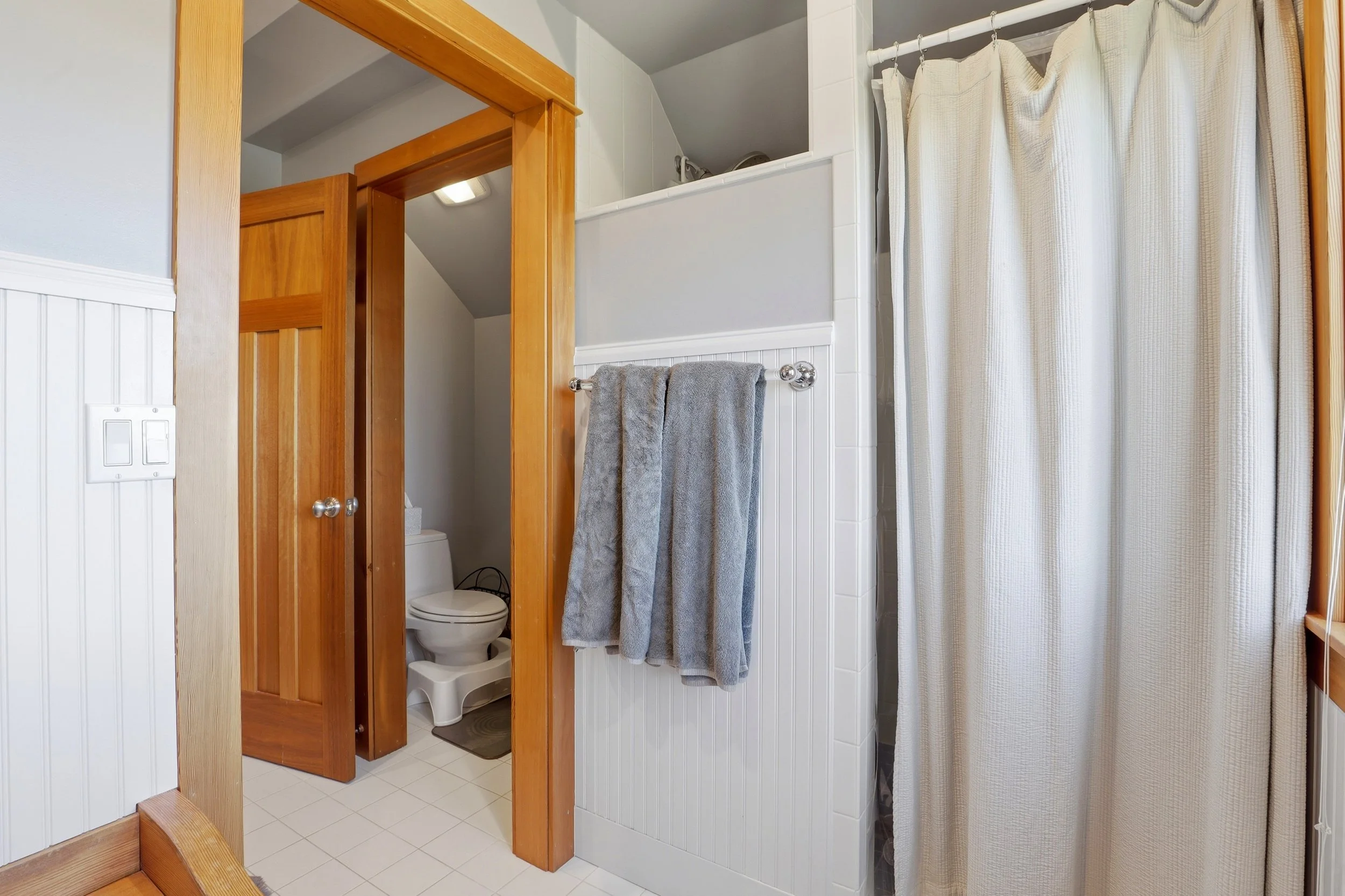 Bathroom with laundry area, including a toilet inside a small closet, a gray towel hanging on a chrome towel rack, a shower with a beige curtain, and wooden trim around the doorway.
