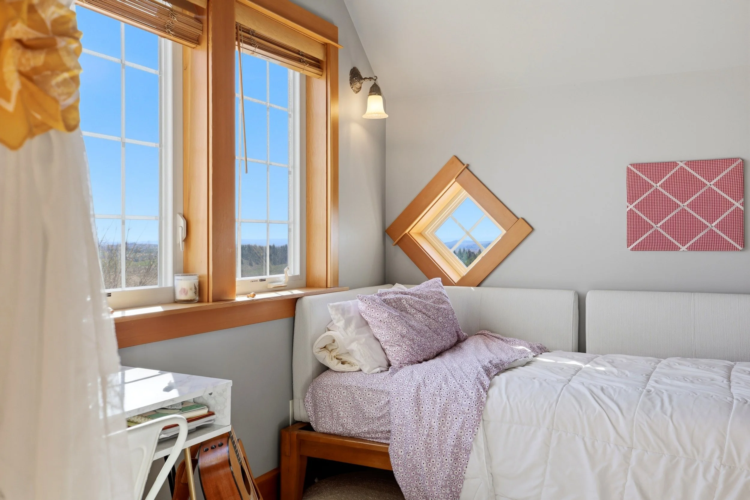 A cozy bedroom with a white bed, purple polka dot bedding, and a single pillow. There are wood-framed windows with a scenic outdoor view of blue sky and landscape. A small white bedside table holds books and a guitar is leaned against it. The walls a