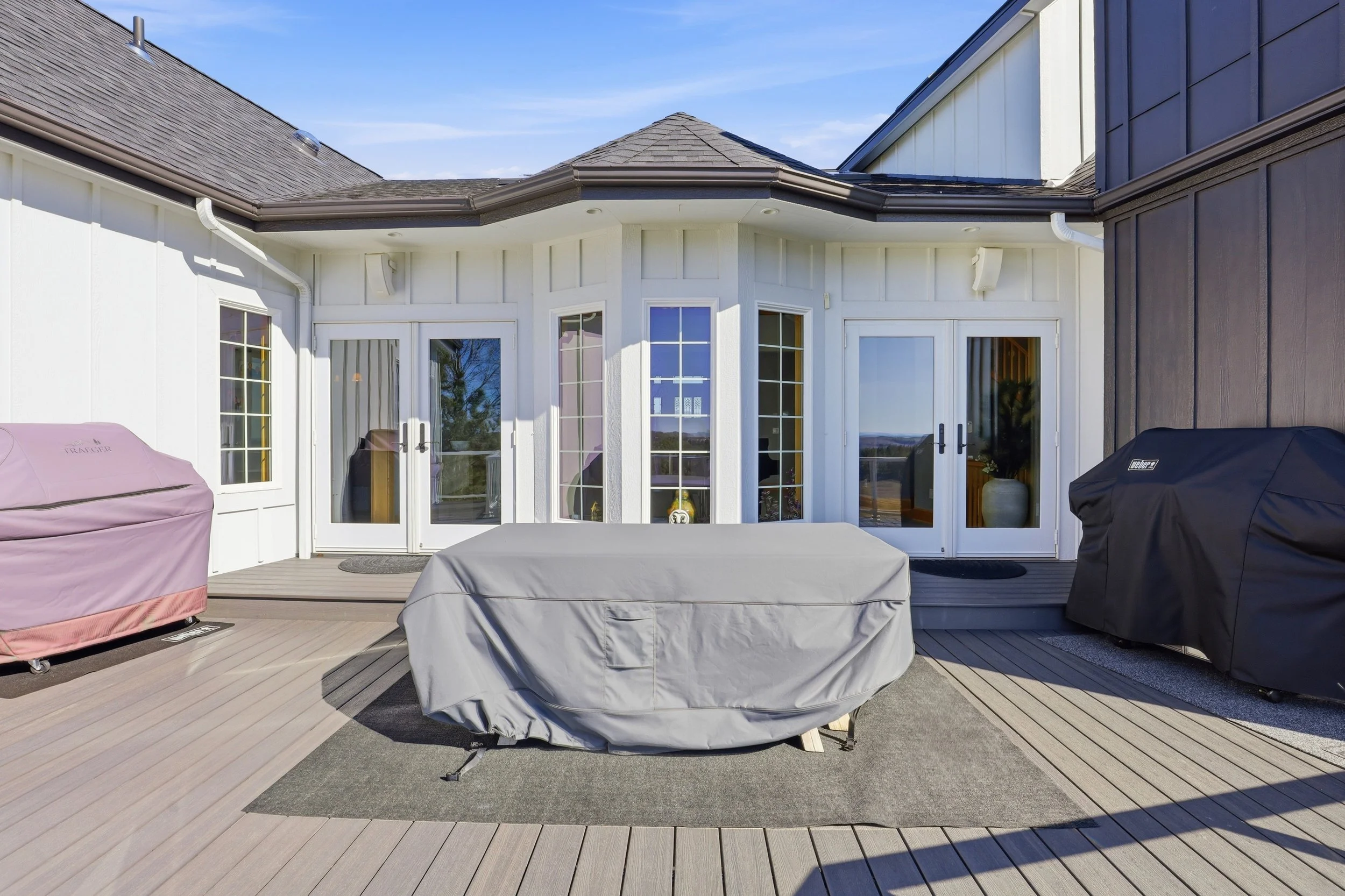 Backyard deck with outdoor furniture covered in protective plastic, two covered grills, and a view of house with white and dark gray siding and multiple windows.