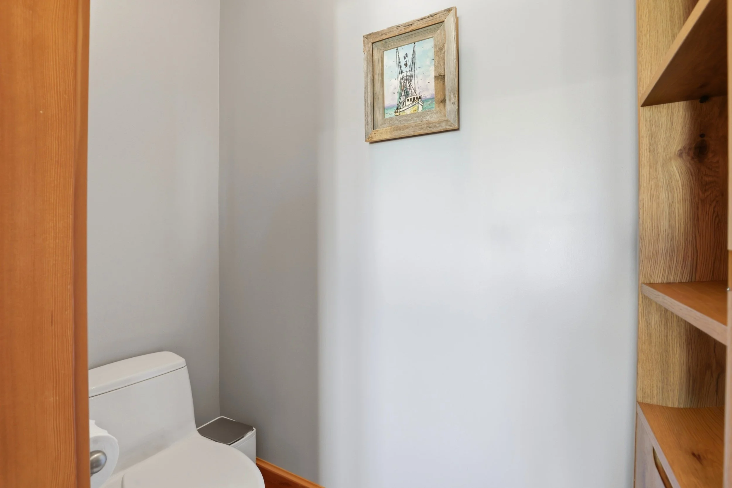 A small bathroom with a white toilet, a white toilet paper holder, beige walls, a wooden frame with a painting of a boat, and a wooden shelf on the right side.