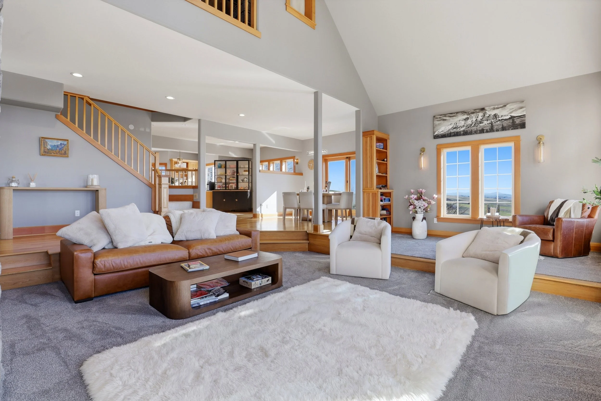 A spacious living room with light gray walls, large windows, and wooden trim. It features white and brown sofas, a wooden coffee table, and a white fluffy rug. There are decor items, books, and plants visible, with a staircase and dining area in the 