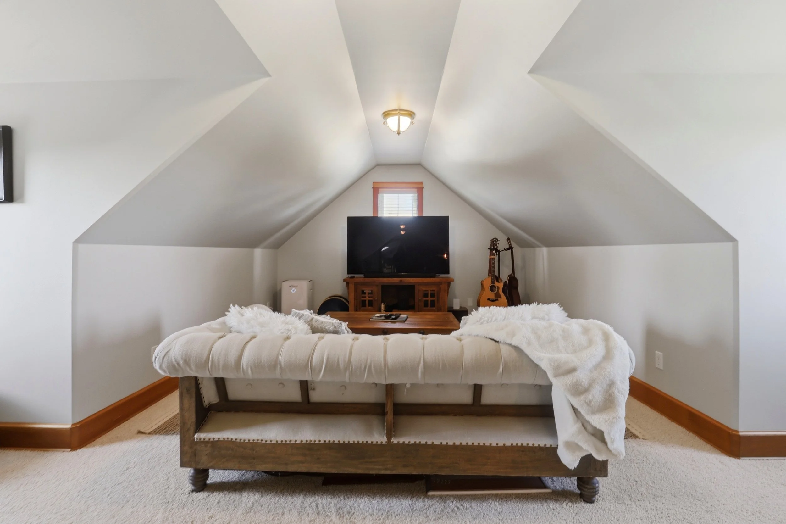 A cozy attic bedroom with a vaulted ceiling, a flat-screen TV on a wooden stand, a guitar leaning against the wall, a bed with white bedding, and a small window with blinds.