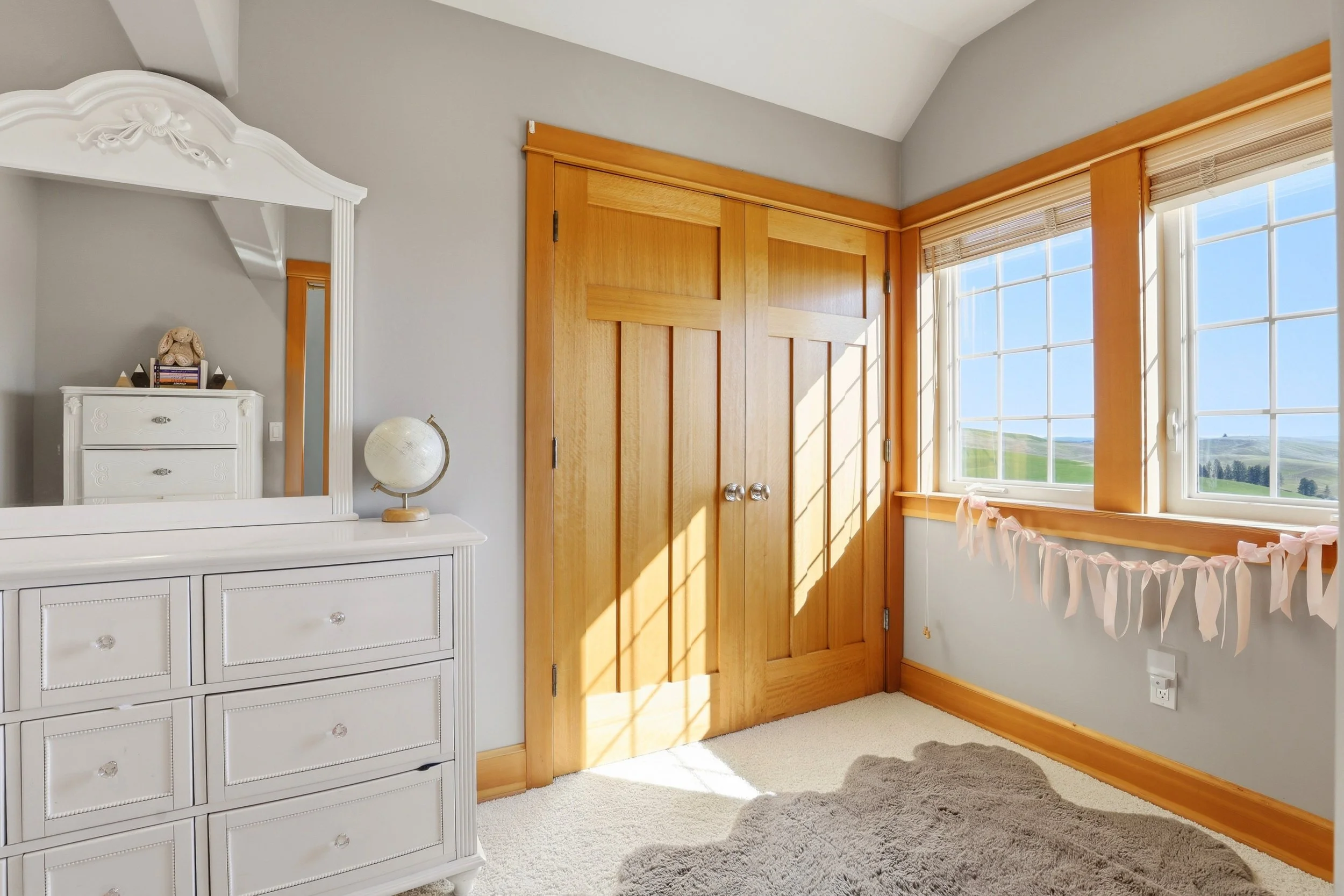 A cozy corner of a room with a white dresser, a globe, a teddy bear, and a mirror hanging above the dresser. Large windows with bamboo blinds and a view of green fields outside. Wooden closet doors and a fluffy gray rug on a beige carpet.