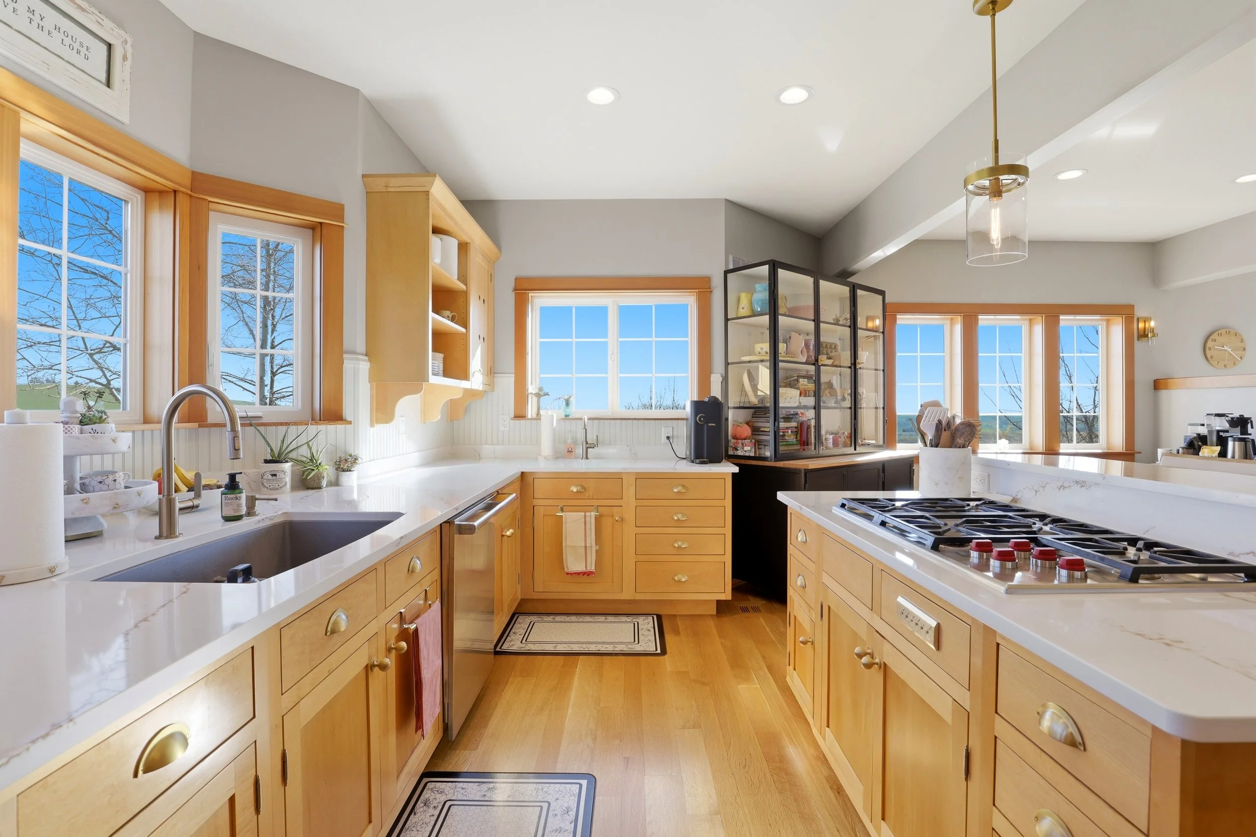 Bright kitchen with wooden cabinets, white countertops, stainless steel sink, stove, large windows showing trees and blue sky, and a glass display cabinet filled with dishes and books.