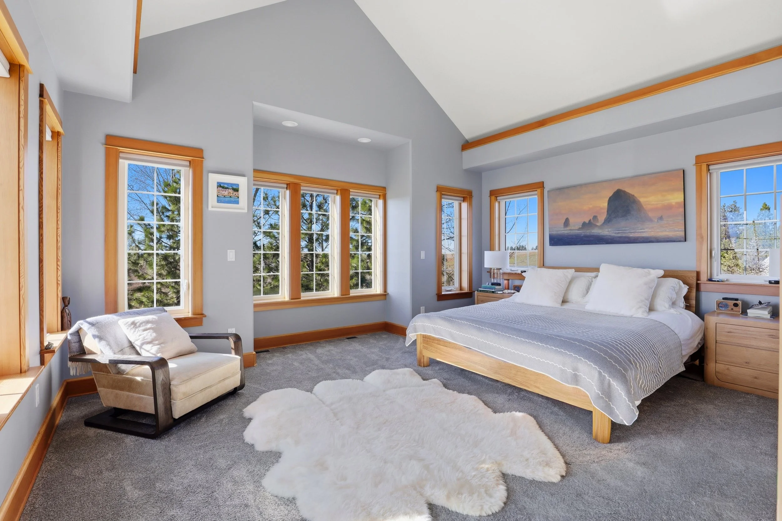 A modern bedroom with multiple windows, a bed with white and gray striped bedding, a white furry rug on gray carpet, a small wooden nightstand with books and a lamp, an armchair with a pillow, and a large landscape painting of rocks and ocean on the 