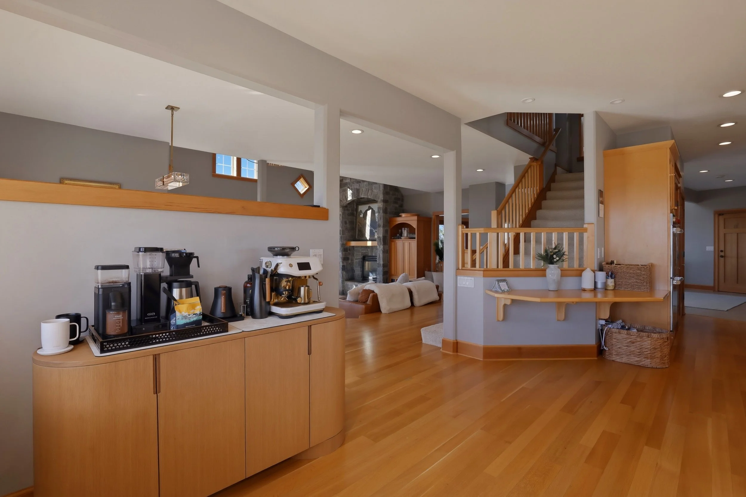 Interior view of a cozy living space with a coffee station on a wooden cabinet, a staircase with wooden railings, and a living area with couches and a stone fireplace. Light-colored walls and hardwood floors add warmth to the room.