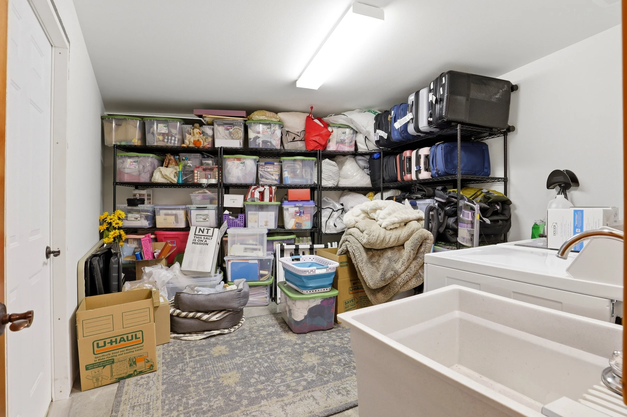 A storage room with black metal shelving filled with plastic bins, suitcases, and miscellaneous items; a U-Haul moving box on the floor, and a laundry sink in the foreground.