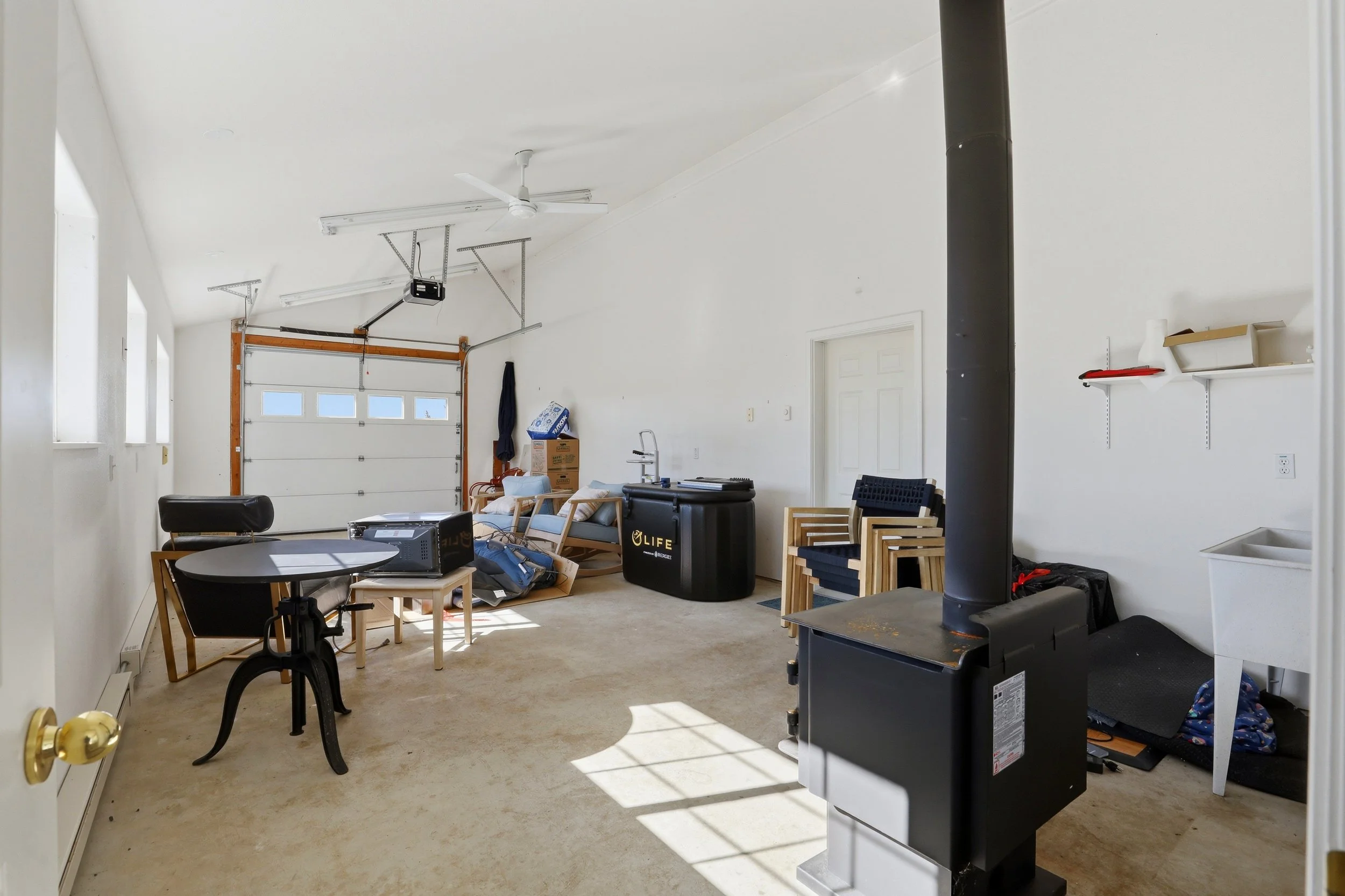 A cluttered garage interior with a white sealed garage door, a black pellet stove, and various furniture and storage boxes used as workspace or storage.