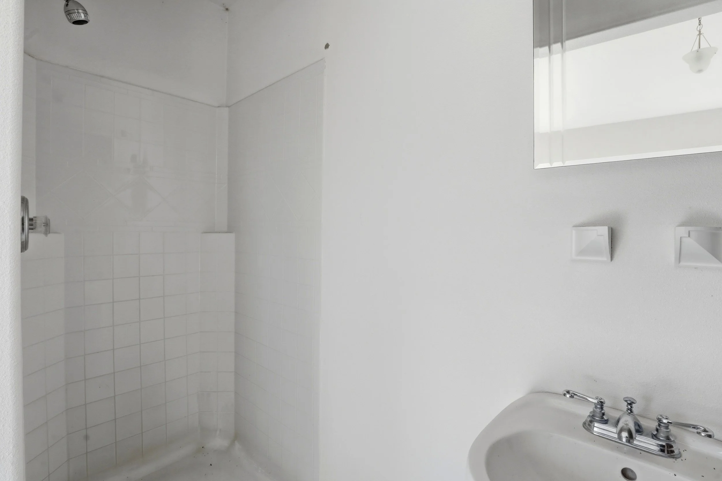 Empty bathroom with white tiled shower area, a mirror on the wall, and a white sink with silver fixtures.
